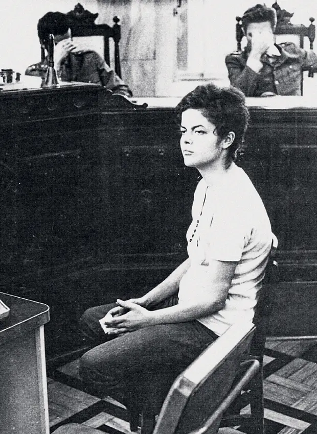 Foto mostra Dilma Rousseff sentada no banco dos réus do Tribunal Militar, em 1970, enquanto dois juízes militares, no plano ao fundo, escondem os rostos com as mãos