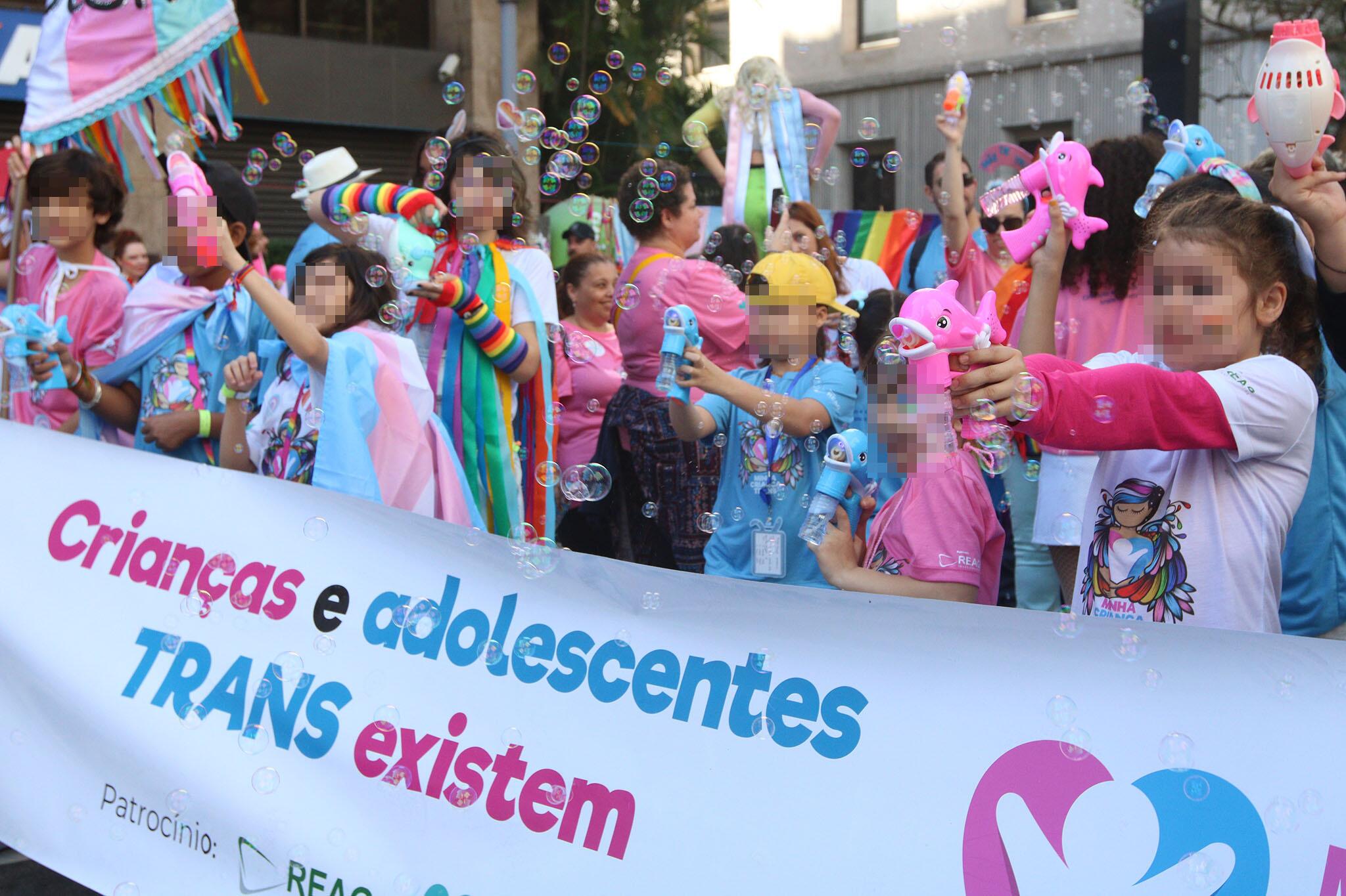 Crianças vestidas com as cores da bandeira trans (rosa claro, azul claro e branco) seguram cartaz com a frase ‘Crianças e adolescentes trans existem’ durante a Parada do Orgulho LGBT de 2023 em São Paulo