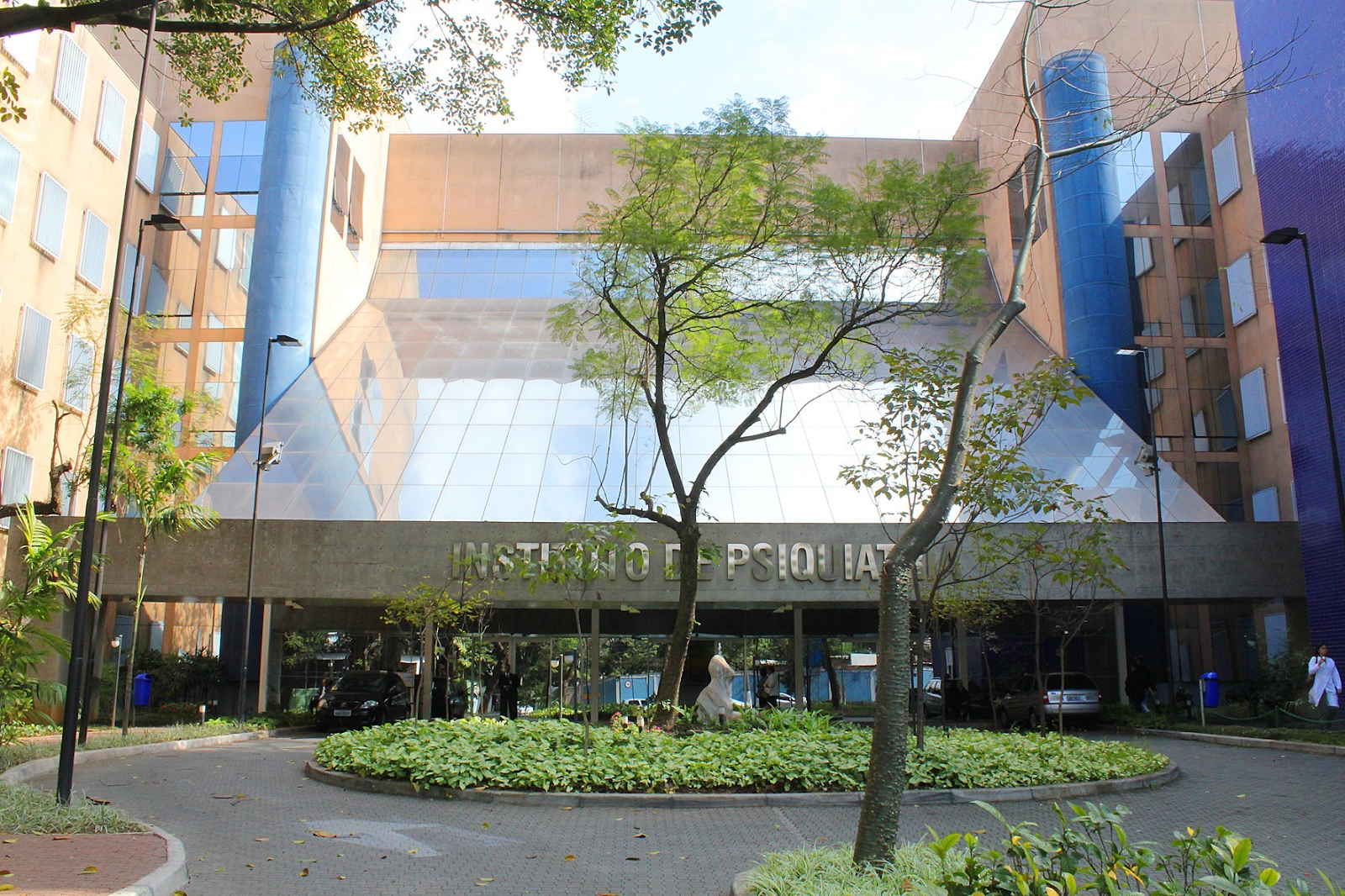 Foto mostra a entrada de um prédio cinza e azul, com árvores, uma estátua e uma passagem para carros, e no canto direito aparece uma mulher vestida de jaleco branco. O local também possui um letreiro informando ser a sede do Instituto de Psiquiatria do Hospital da Clínicas da USP, em São Paulo