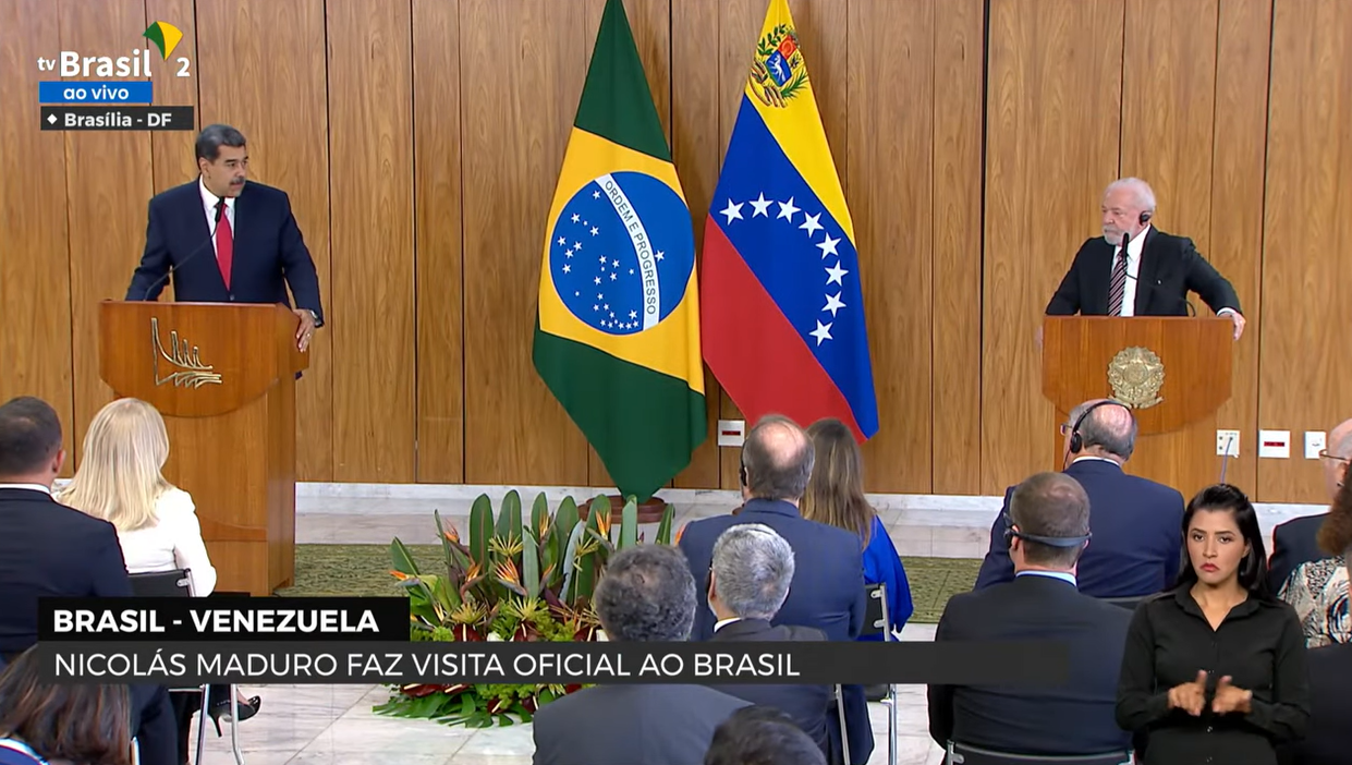 Imagem mostra Lula, à direita, e Maduro, à esquerda. Ambos vestem ternos escuros, estão atrás de pulpitos de madeira e são observados por uma plateia.  Entre eles aparecem as bandeiras do Brasil e da Venezuela.