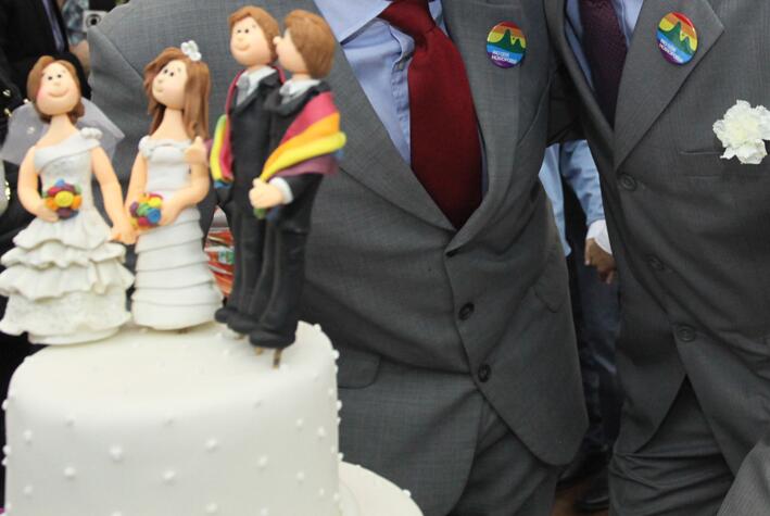 Em primeiro plano, há um bolo de casamento com bonecos que representam dois casais homoafetivos. Ao fundo, duas pessoas de terno se abraçam e usam broches com a bandeira LGBTQIA+