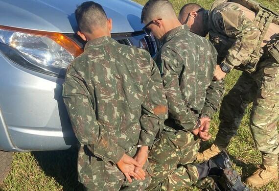 Dois homens fardados ajoelhados em frente a um carro são algemados por policial militar