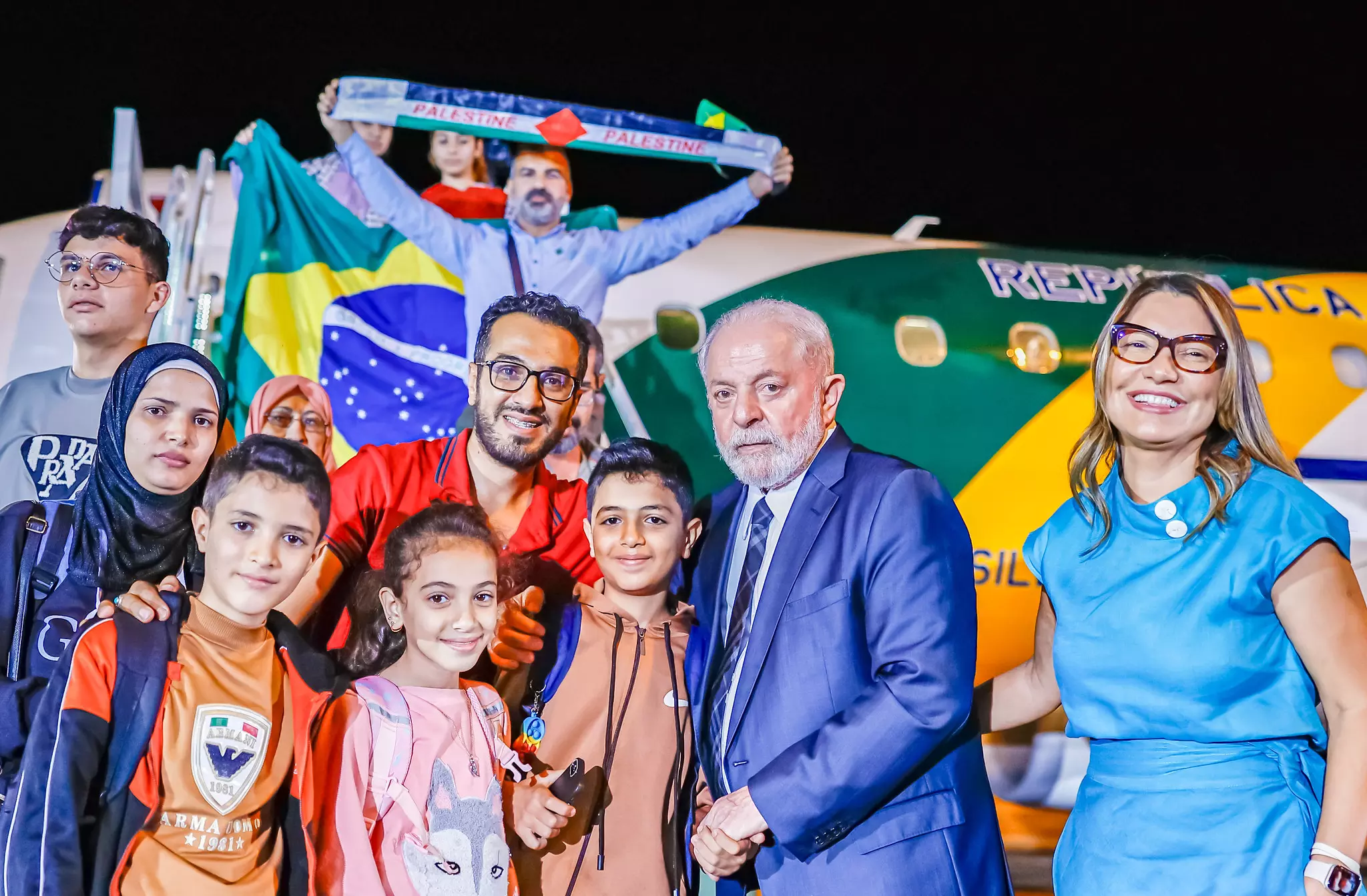 O presidente Lula e a primeira-dama Rosângela da Silva aparecem ao lado de crianças e adultos vindos da Faixa de Gaza. Ao fundo, aeronave da Força Aérea Brasileira responsável por trazer grupo ao Brasil