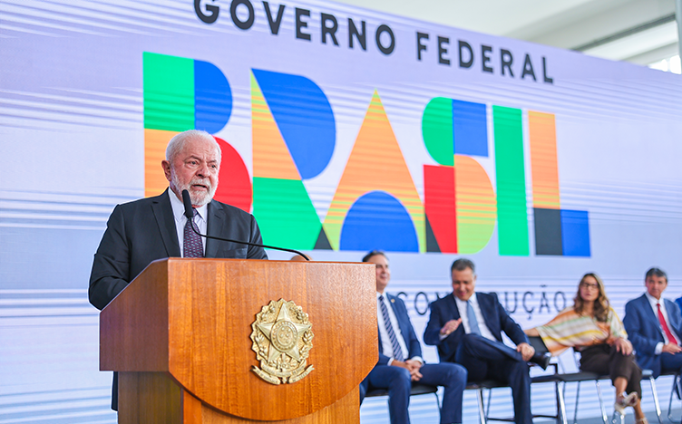 Em primeiro plano, Lula discursa em púlpito; ao fundo, ministros estão sentados em cadeiras e há painel com logo do governo federal.