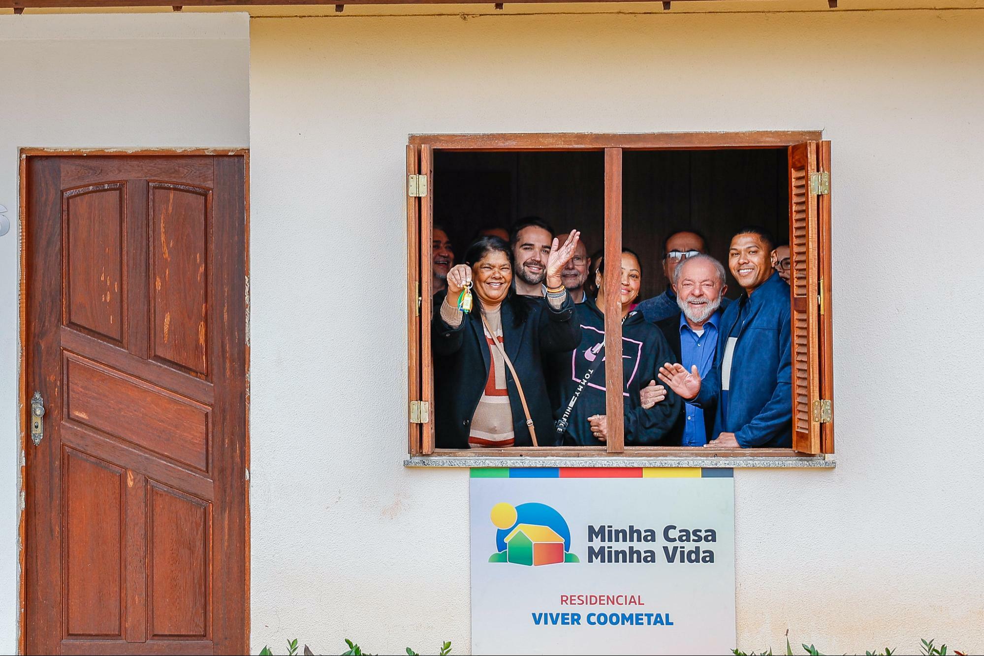 Fachada de casa do projeto Minha Casa, Minha Vida. Na janela, o presidente Lula e o governador do Rio Grande do Sul, Eduardo Leite, sorriem ao lado de um grupo de pessoas.
