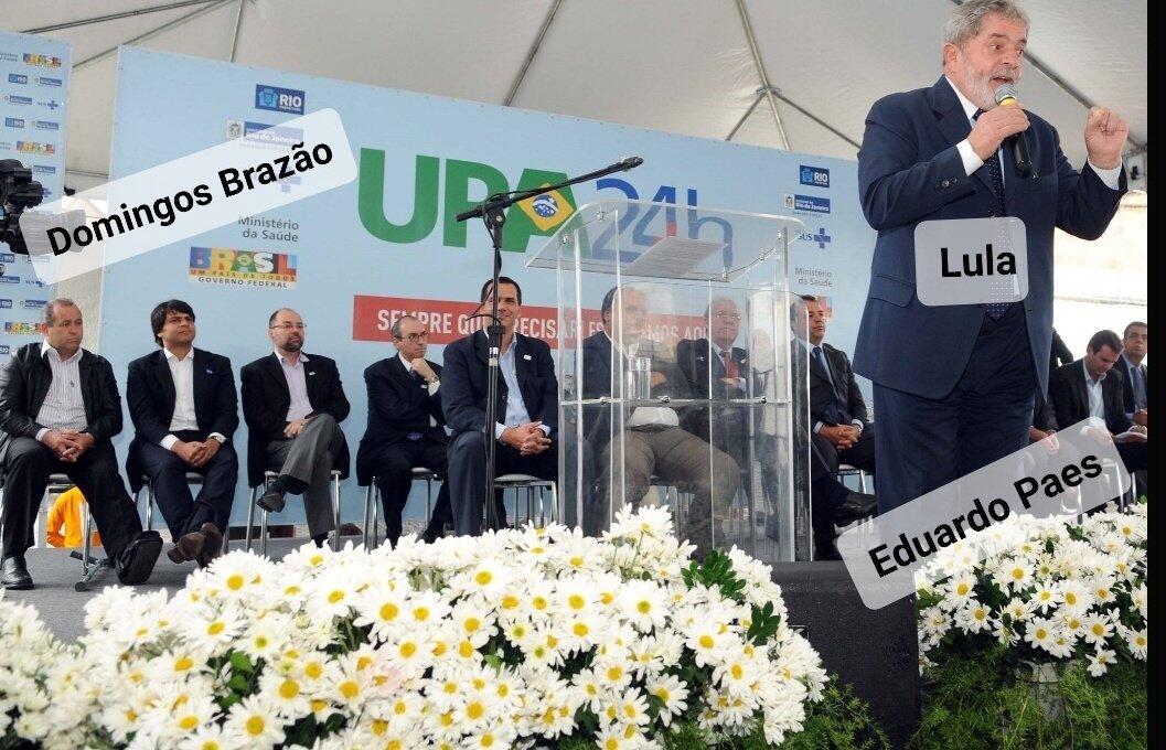 Foto mostra Lula discursando. Ao fundo, Domingos Brazão está sentado junto de outras personalidades da política carioca.
