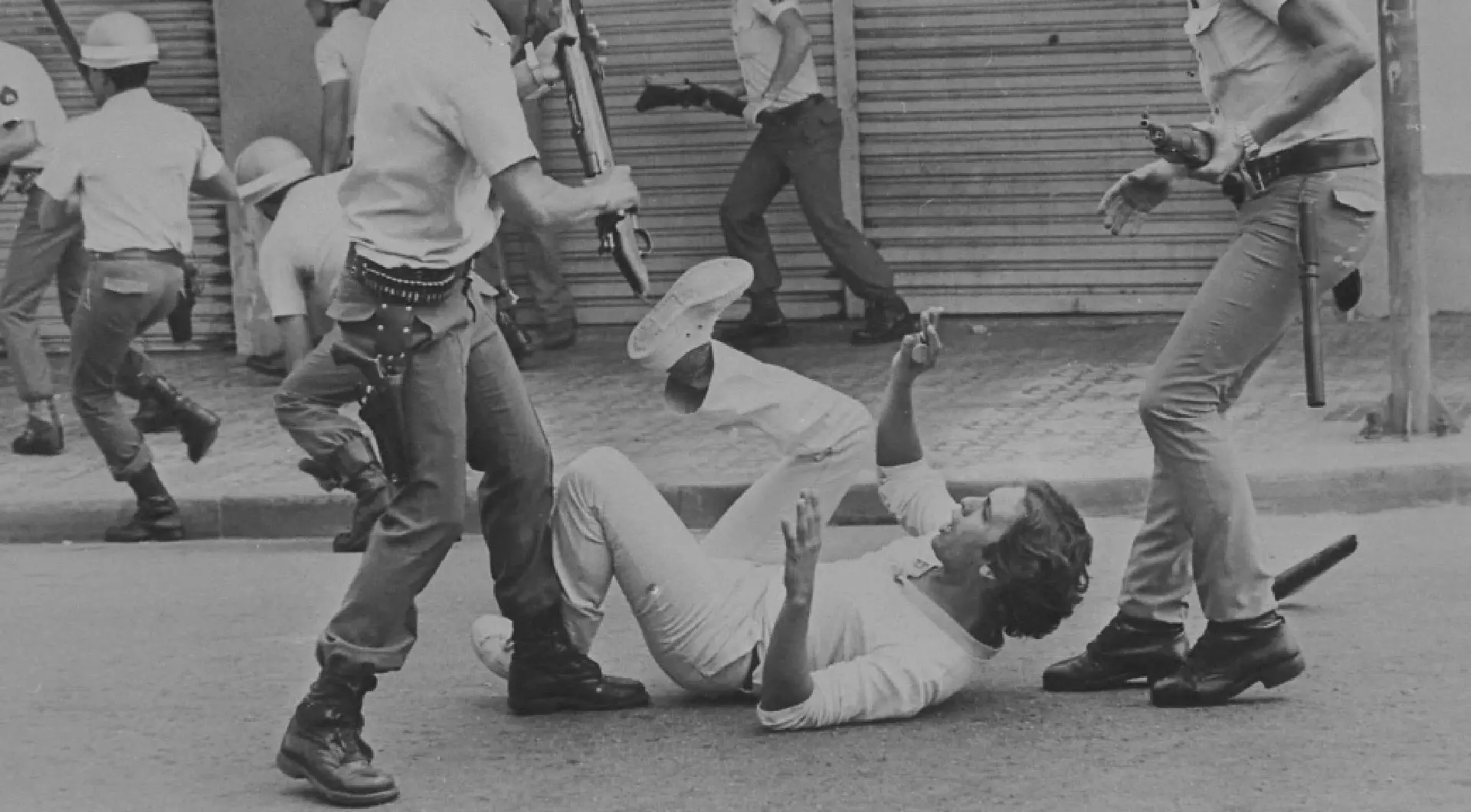 Homem caído no chão está com os braços estendidos para se proteger do golpe de um militar que está prestes a agredi-lo