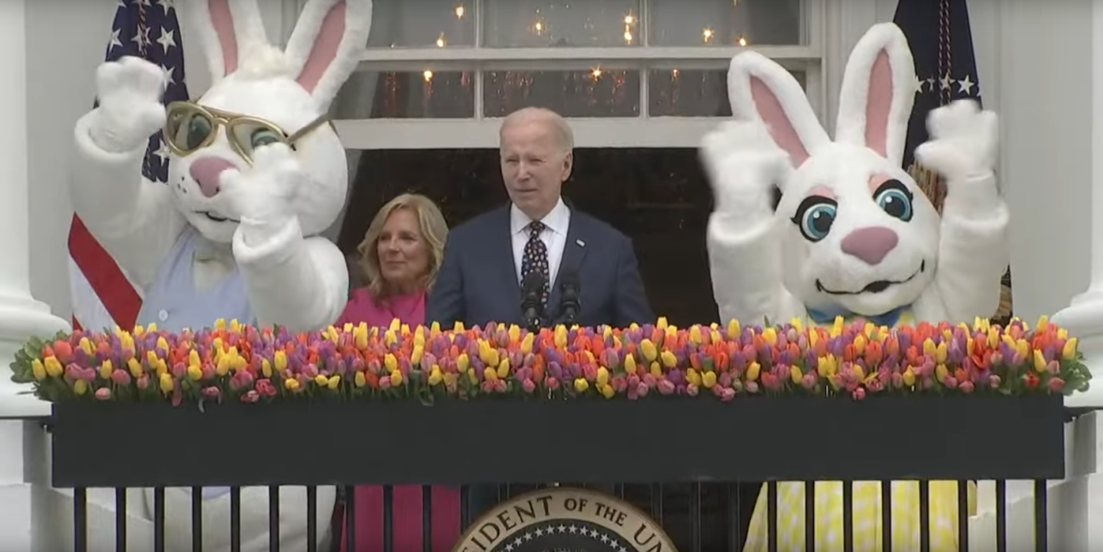 Biden discursa enquanto duas pessoas vestidas de coelhos da Páscoa, ao seu lado, acenam para o público