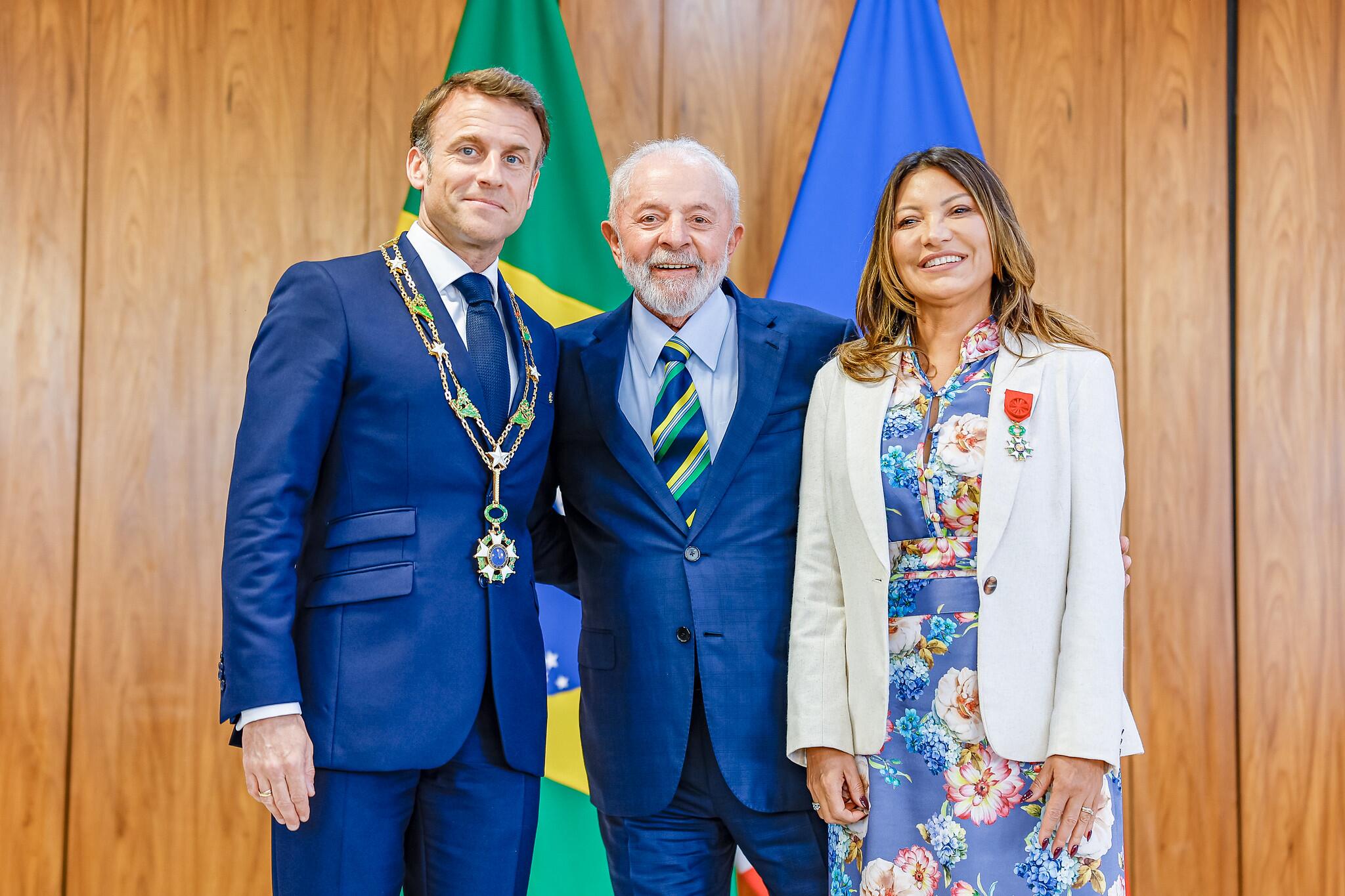 Lula, de terno azul, está no centro da imagem, abraçado a Emmanuel Macron, também de terno azulado, e Janja, que usa um blazer branco com um broche vermelho