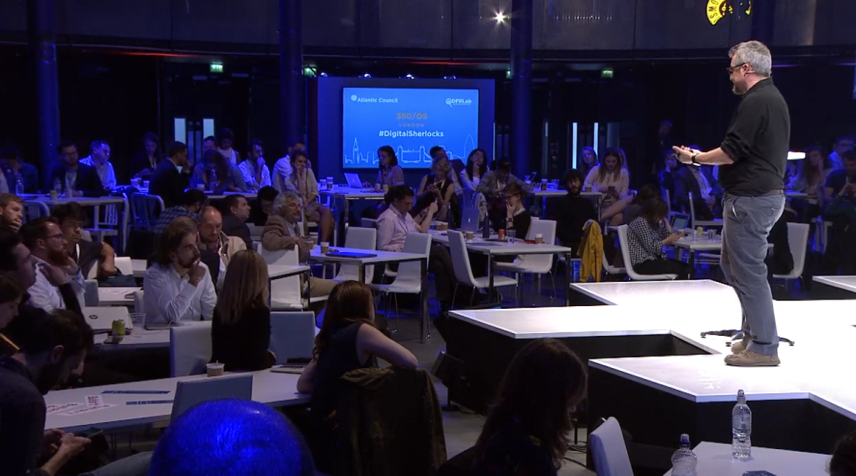 Homem branco, grisalho e de óculos, faz apresentação sobre um palco branco para plateia. Participantes têm laptops sobre as mesas e estão sob uma luz azulada. Ao fundo, há um telão com fundo azul em que se lê o nome do evento e a hashtag #digitalsherlocks