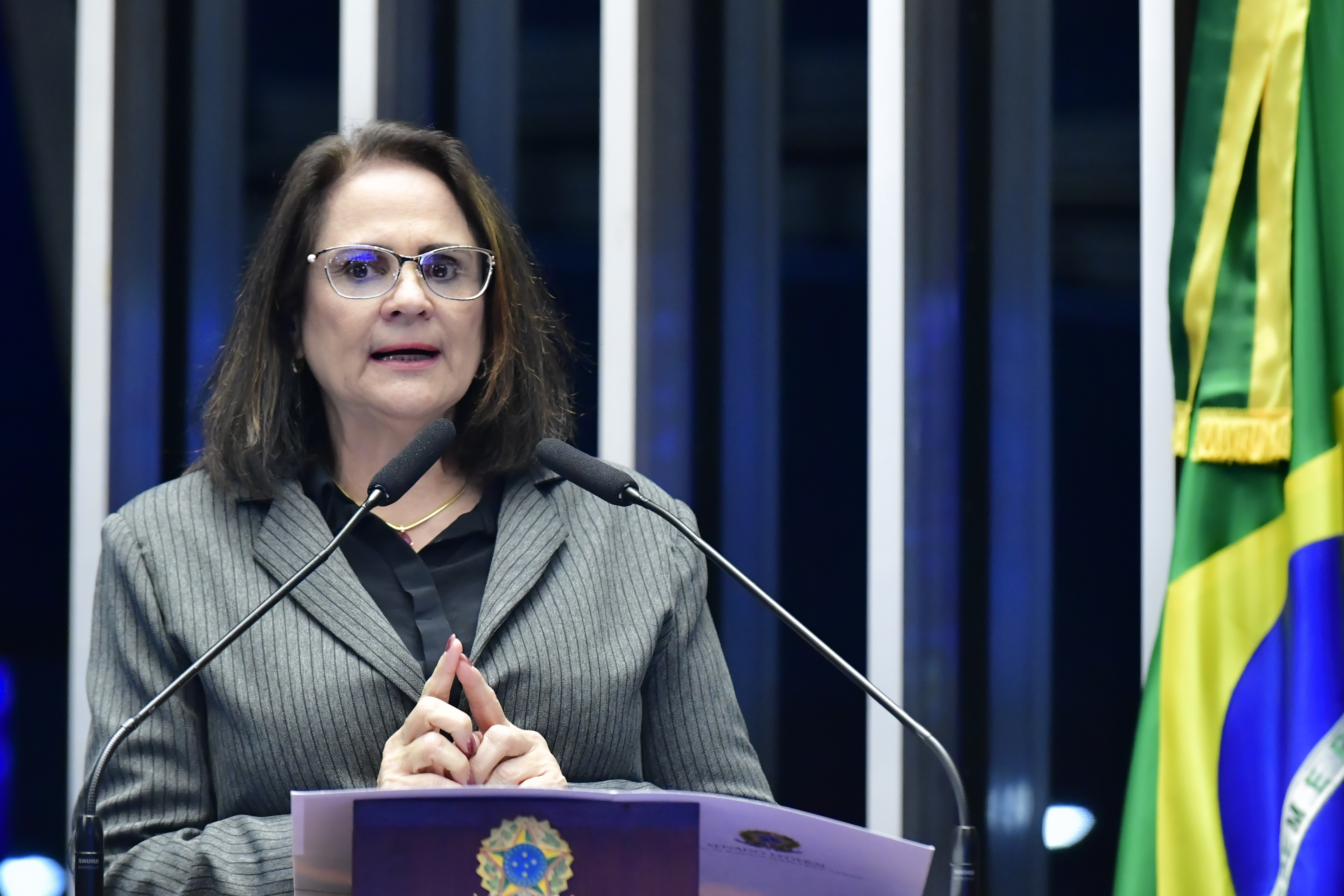 Senadora Damares, mulher branca, de óculos e com cabelos escuros na altura do ombro, usa terno cinza listrado sobre camisa preta. Ela discursa na tribuna, onde se vê o brasão da República, ao lado da bandeira do Brasil.