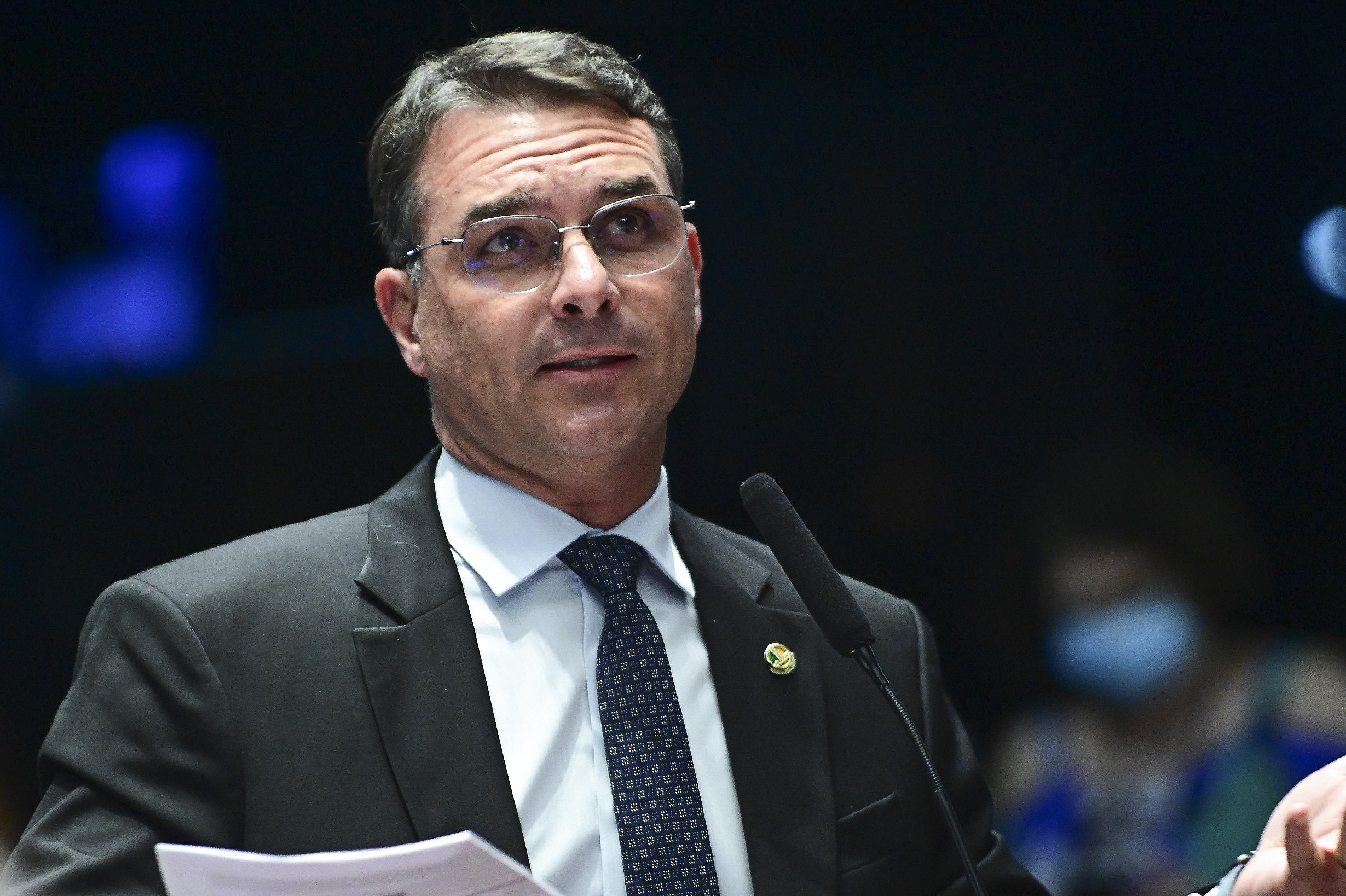 Flávio Bolsonaro é um homem branco de meia idade, com cabelos castanhos claros e óculos de aros finos. Ele veste terno escuro sobre camisa clara e gravata azul marinho com estampa discreta.