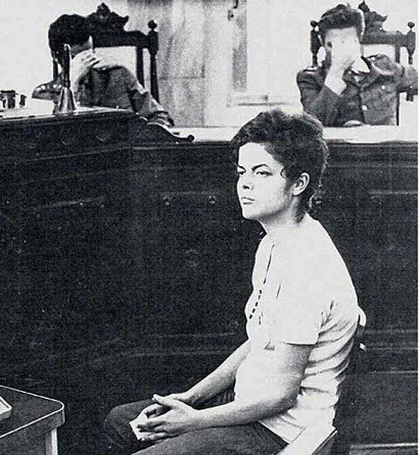 Foto em preto e branco. Dilma, uma mulher branca de cabelo curto e com feição séria vestindo uma camiseta branca e calça preta. Ao fundo dois militares escondem os rostos com as mãos.