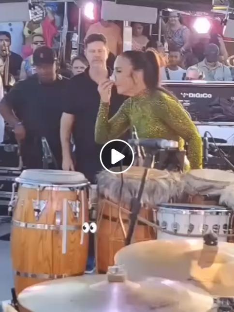 Imagem mostra Ivete Sangalo, mulher branca e com roupa verde brilhante, em cima de um trio elétrico com diversas pessoas e instrumentos musicais. Ela segura um algodão próximo a seu nariz.