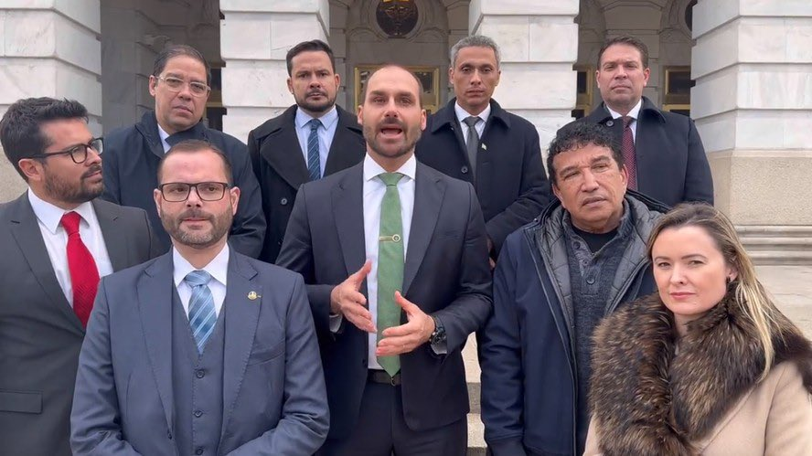 Eduardo Bolsonaro aparece ao centro, discursando em frente a um edifício de arquitetura branca ao lado de uma comitiva de parlamentares e apoiadores brasileiros. Todos vestem roupas formais ou casacos de frio.
