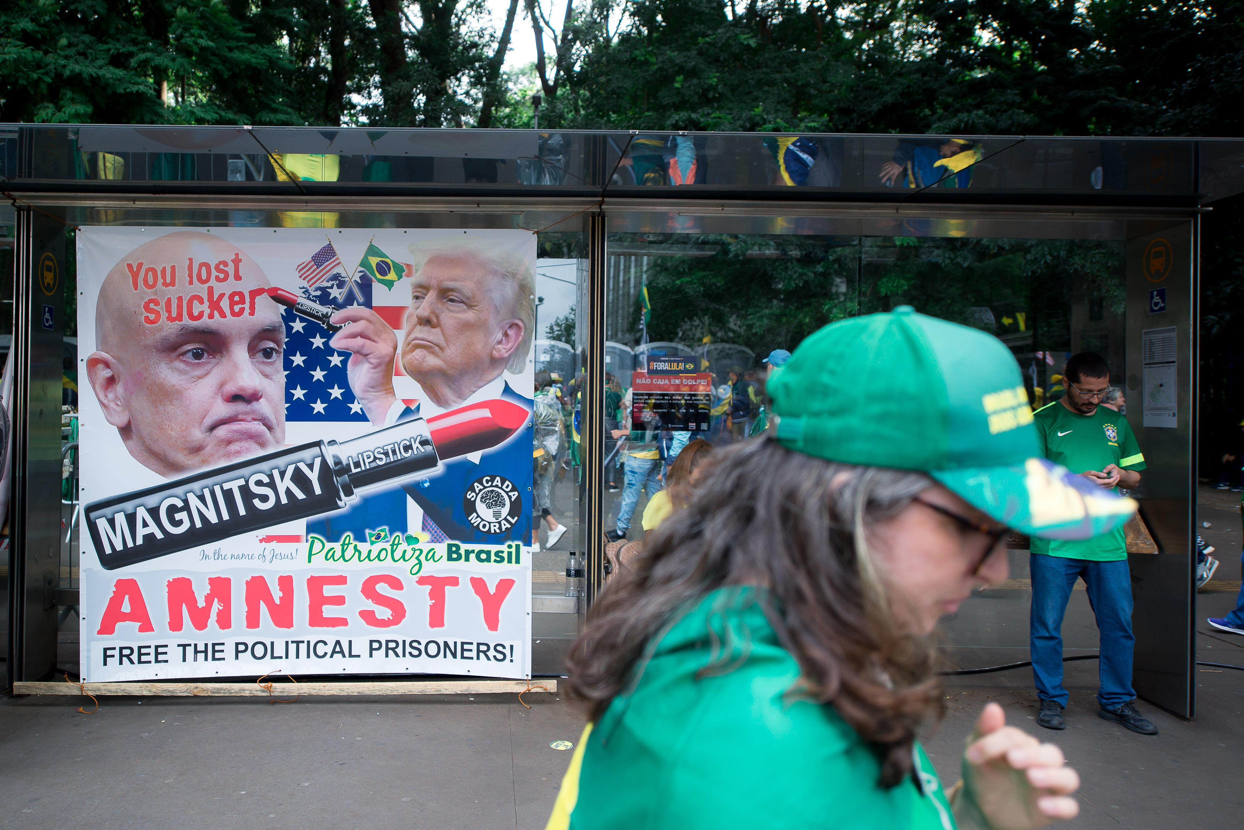 Manifestante de boné verde e amarelo circula diante de um cartaz com montagem de Donald Trump escrevendo ‘You lost sucker’ com batom na testa do ministro Alexandre de Moraes. No centro do banner, há um batom onde se lê ‘Magnitsky lipstick’. Abaixo está escrito ‘Amnesty – Free the political prisoners!’. O cartaz é assinado por ‘Patriotiza Brasil’.
