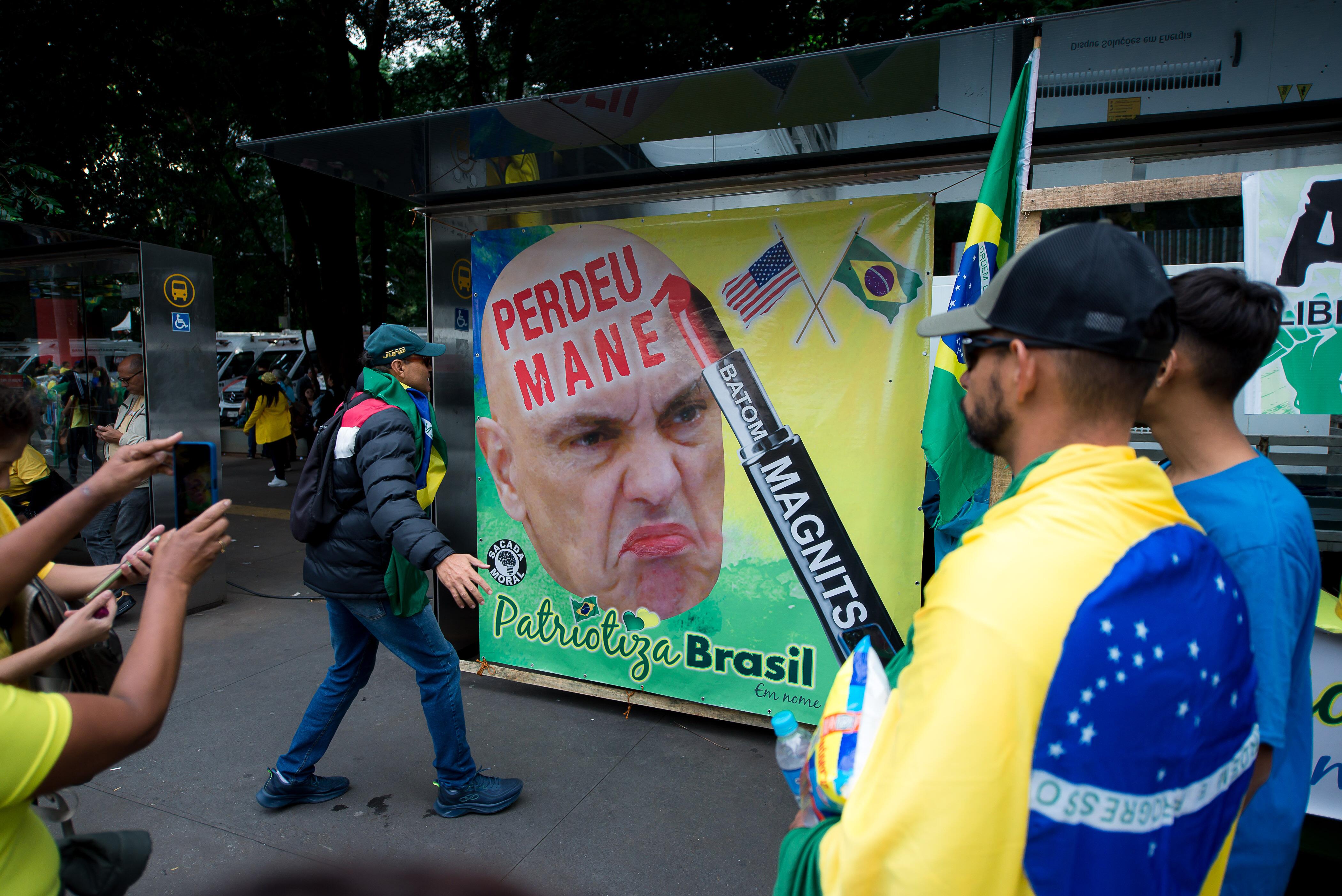 Manifestantes com roupas nas cores da bandeira do Brasil observam cartaz com a imagem caricaturada do ministro Alexandre de Moraes, com a inscrição ‘Perdeu Mané’ em vermelho na testa. A arte inclui batom com a palavra ‘Magnits’, bandeiras dos EUA e do Brasil, e a assinatura do grupo ‘Patriotiza Brasil’.