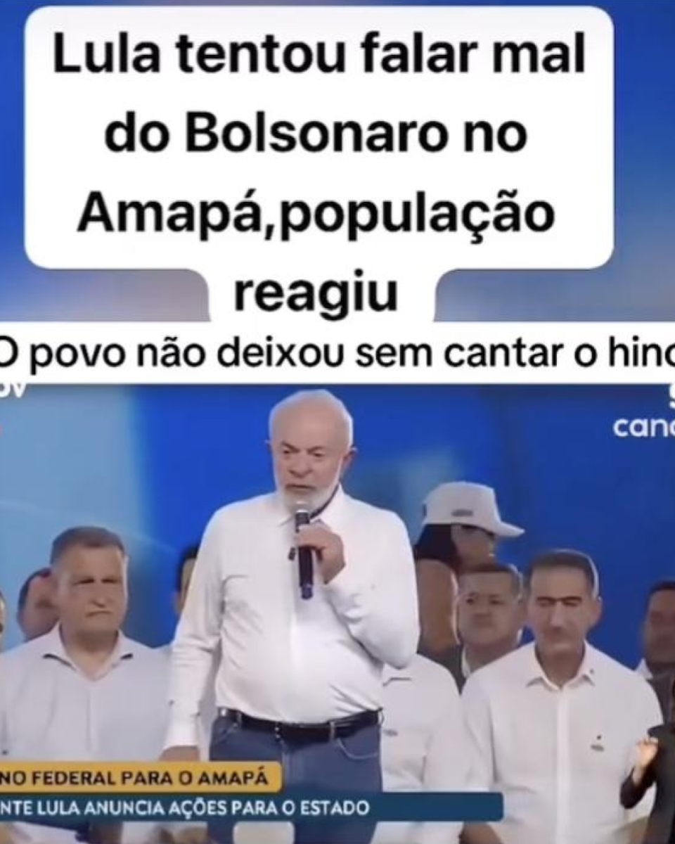 Print de um vídeo publicado no Instagram mostra um evento público com o presidente Lula (PT) em destaque, falando ao microfone. Ele é um homem branco, de barba branca e cabelos brancos, que veste uma camisa social branca de mangas compridas e calça jeans azul. Ao fundo, há diversas pessoas, todas vestidas de branco, algumas usando boné. No topo da imagem, há a sobreposição de um texto em letras pretas sobre fundo branco que diz: ‘Lula tentou falar mal do Bolsonaro no Amapá, população reagiu’. Abaixo, outro texto diz: ‘O povo não deixou sem cantar o hino’.