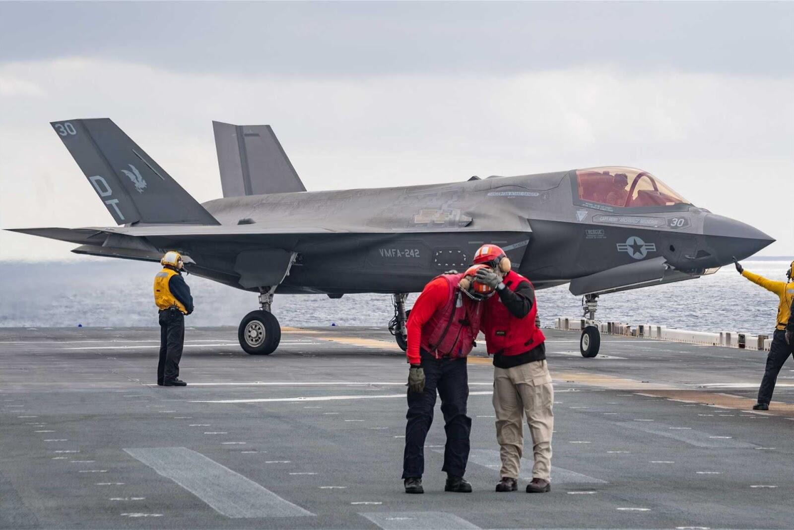 Um avião de caça F-35 cinza escuro está estacionado no convés de um porta-aviões. Vários membros da equipe estão ao redor do avião, incluindo dois em primeiro plano com coletes vermelhos e capacetes, um deles com as mãos na cabeça do outro. Ao fundo, o oceano e o céu nublado são visíveis.
