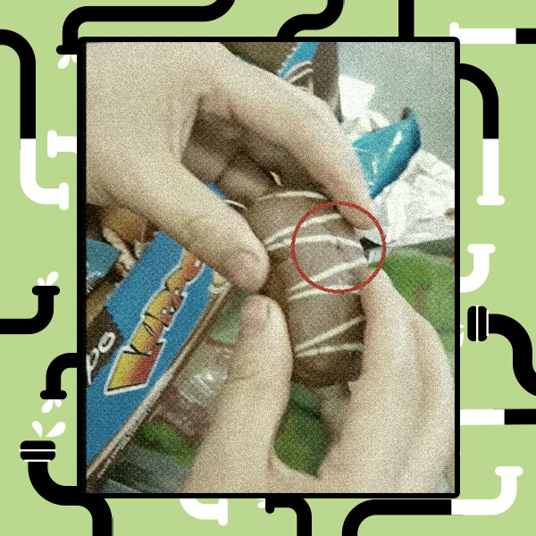 Uma foto de perto, tirada de cima, mostra duas mãos segurando um bolo ou doce em formato de cilindro com cobertura de chocolate e linhas brancas. Um círculo vermelho é desenhado em torno de um pequeno ponto no bolo. Há embalagens de comida embaçadas no fundo.