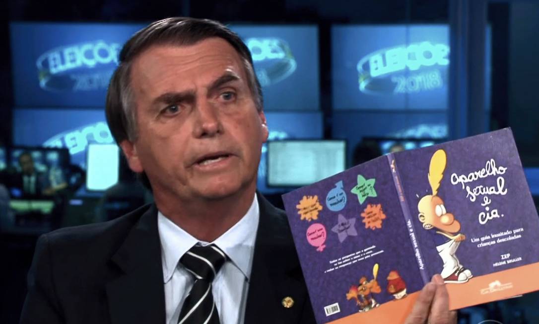 Jair Bolsonaro, vestindo terno escuro, camisa branca e gravata listrada, aparece sentado em um estúdio de TV durante as eleições de 2018. Ele segura aberto, voltado para a câmera, o livro infantil ‘Aparelho Sexual e Cia.: Um guia inusitado para crianças descoladas’, com capa roxa e ilustrações coloridas. Ao fundo, há monitores com o logotipo ‘ELEIÇÕES 2018’, indicando o contexto de uma entrevista ou debate televisivo.