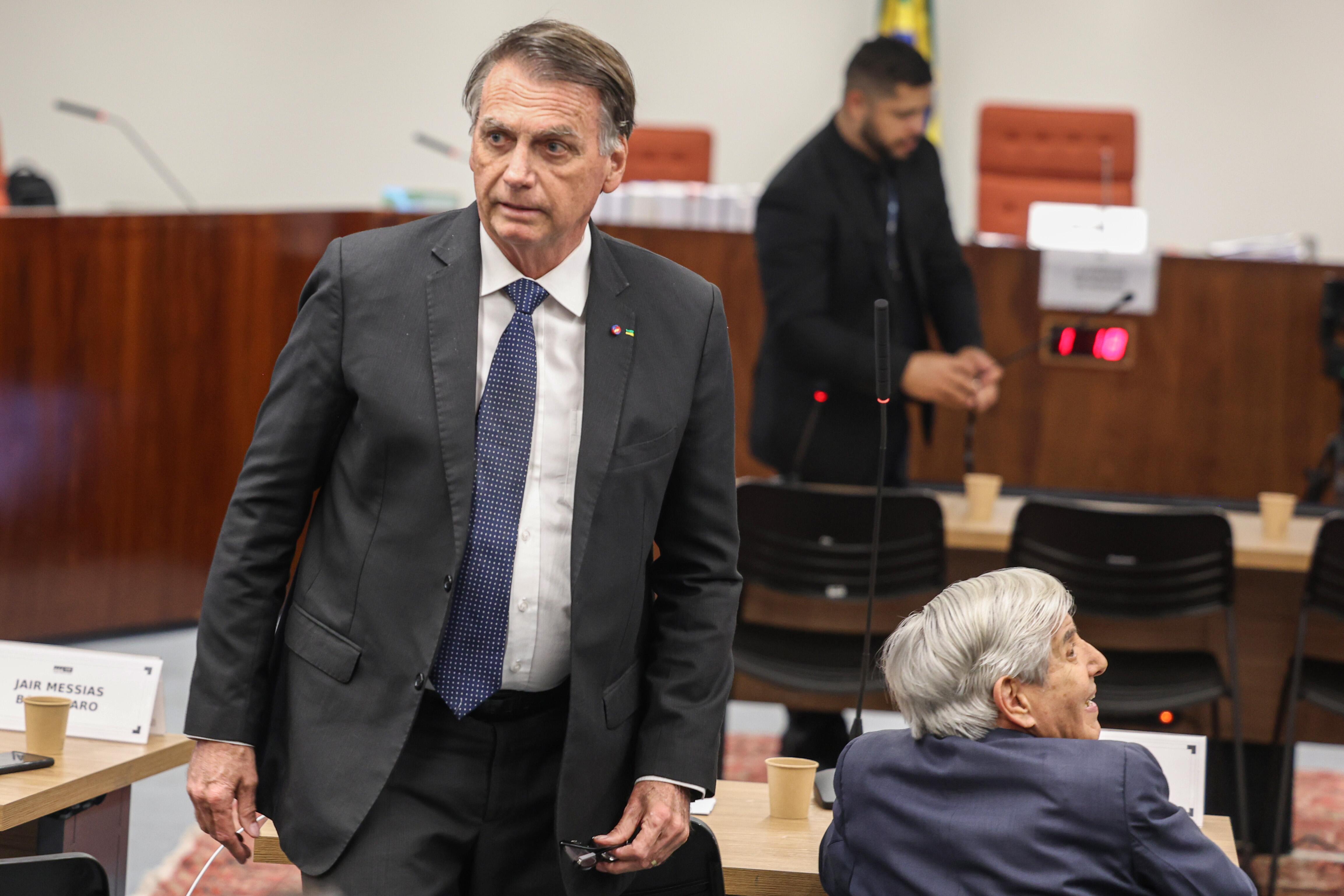 Jair Bolsonaro, de terno escuro e gravata azul, está em pé em uma sala de audiências. Ele segura um par de óculos na mão direita e olha para o lado com expressão séria. À sua frente, um homem idoso de cabelos grisalhos está sentado e virado para trás, olhando para Bolsonaro. Ao fundo, um homem de terno preto interage com algo sobre uma mesa. O ambiente é formal, com mesas, cadeiras e microfones, sugerindo um tribunal ou audiência oficial.