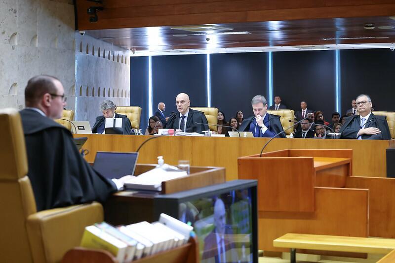 Ministros do Supremo Tribunal Federal estão reunidos no plenário durante uma sessão. Sentados em cadeiras amarelas atrás de uma bancada de madeira, os ministros usam togas e estão voltados para um colega que aparece de costas, também togado, falando de sua bancada. Ao fundo, outras pessoas acompanham a sessão, sentadas em uma área reservada ao público. O ambiente é formal, com iluminação artificial e decoração em mármore e madeira.