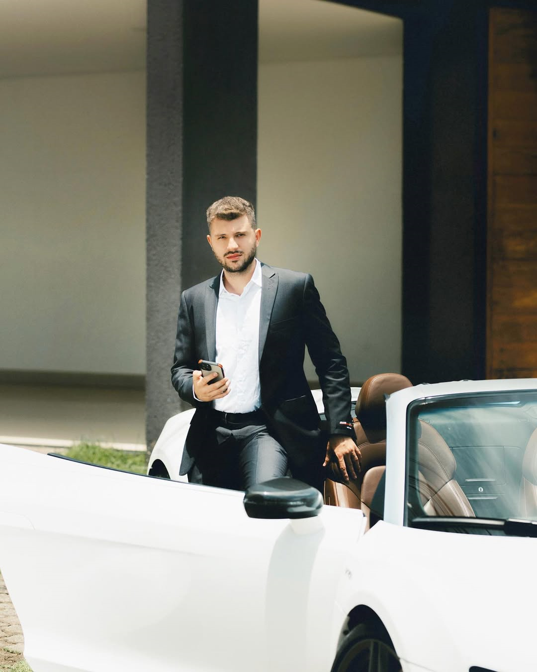 Homem jovem de barba curta, vestido com terno preto e camisa branca, segura um celular enquanto sai de um carro esportivo branco conversível, estacionado em frente a uma casa moderna com fachada minimalista.