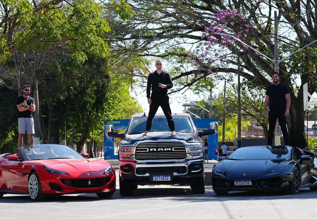 Três homens posam em cima de carros de luxo estacionados em uma rua arborizada: à esquerda, um homem em pé sobre uma Ferrari vermelha conversível; ao centro, um homem descalço de roupa preta em pé no capô de uma caminhonete RAM; à direita, outro homem em pé sobre um Lamborghini preto.