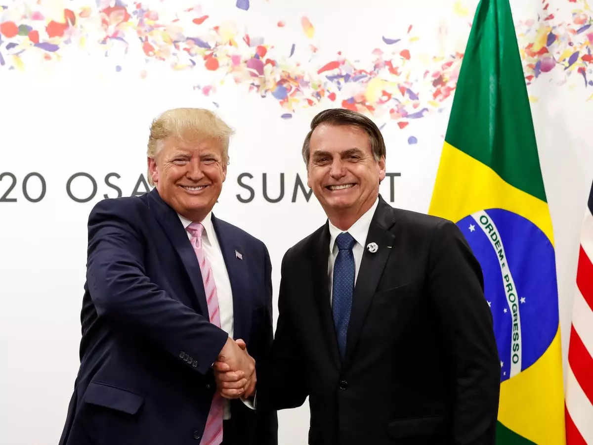 Donald Trump e Jair Bolsonaro apertam as mãos sorridentes durante o encontro do G20 em Osaka, em 2019. Ao fundo, aparecem as bandeiras dos Estados Unidos e do Brasil, e parte da inscrição “G20 Osaka Summit” decorada com confetes coloridos no topo.