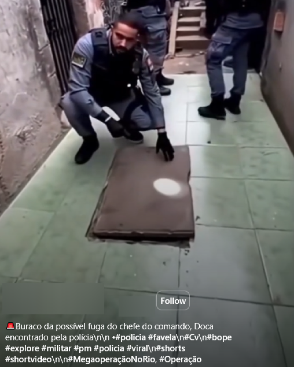 A imagem mostra um grupo de homens vestindo uniformes escuros com a palavra ‘Polícia’ escrita nas costas, em um beco estreito entre construções de alvenaria. Um deles, de pele clara e cabelo curto escuro, está agachado e usa luvas pretas, apontando para uma tampa retangular no chão. Outros policiais permanecem em pé ao fundo, observando a cena.