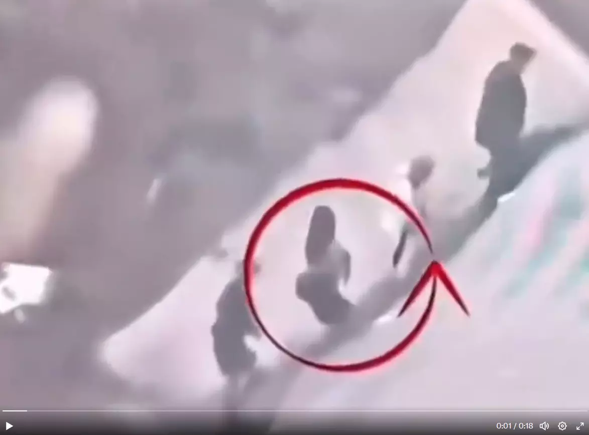 Uma captura de tela de um vídeo, com baixa resolução e aparência enevoada, mostra uma cena em uma superfície clara e inclinada. Um grande círculo vermelho com uma seta, sobreposto à imagem, aponta para uma figura central vestida com roupas escuras, que está em uma posição inclinada para trás. Ao fundo, outras figuras humanas desfocadas estão presentes na mesma superfície.