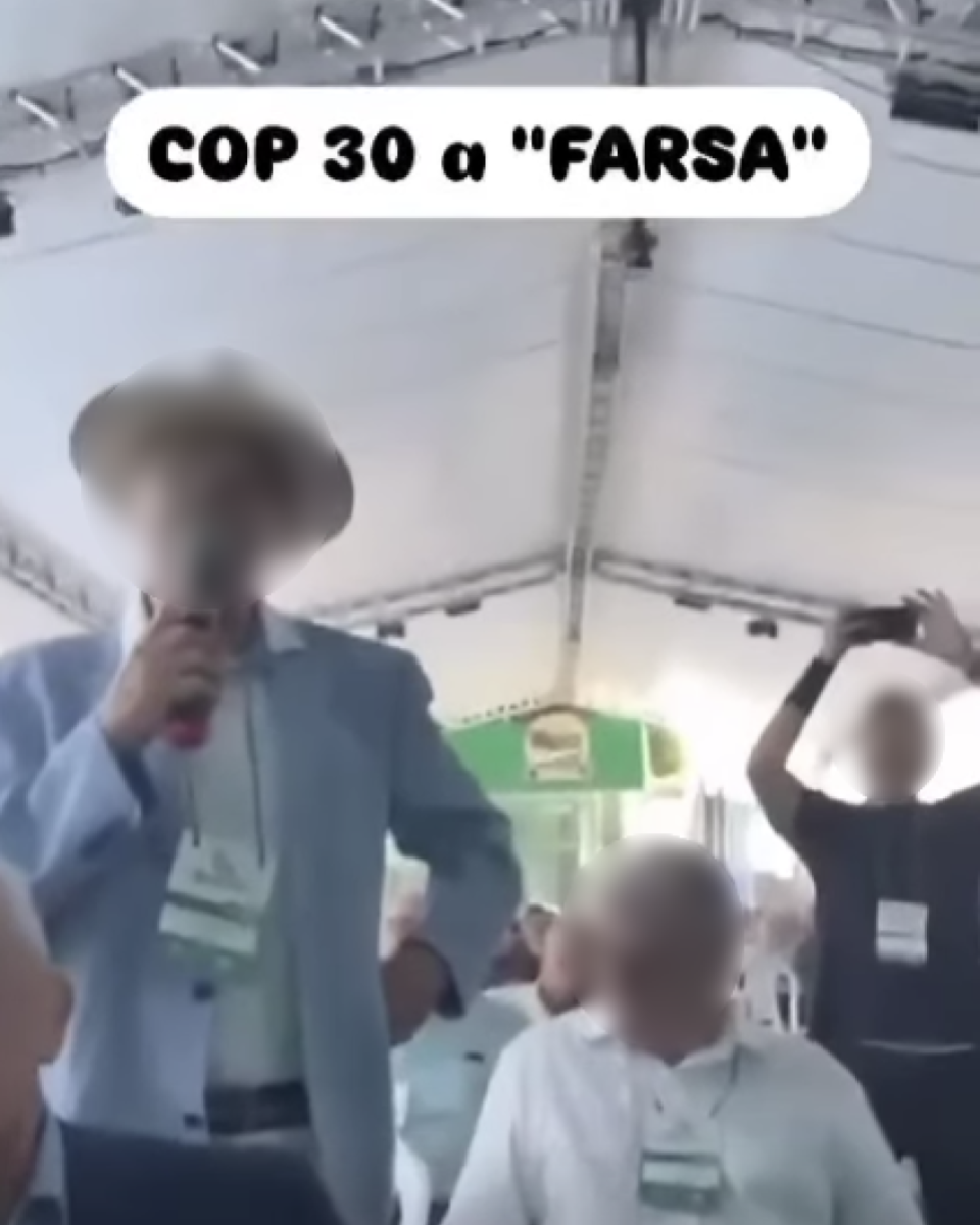 A imagem mostra um homem em pé, segurando um microfone e falando sob uma tenda branca. Ele veste um paletó azul-claro, camisa branca e chapéu bege. À frente dele, há pessoas sentadas, também vestindo roupas claras e crachás pendurados no pescoço. No fundo, outra pessoa está de pé, levantando os braços e segurando um celular para gravar ou tirar fotos. Na parte superior da imagem há um texto em letras pretas dentro de uma faixa branca que diz: 'COP 30 a ‘FARSA’'.