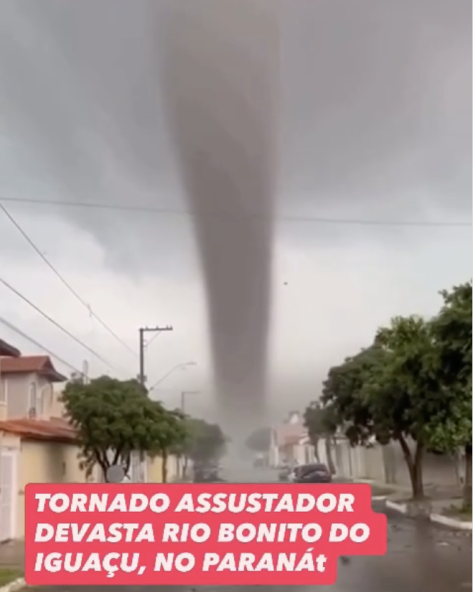 Print de publicação no Instagram de uma cena gerada por IA (inteligência artificial) que mostra uma rua residencial com casas de telhados alaranjados e árvores. No centro, há um grande funil de tornado que se estende do céu nublado até o solo. O asfalto apresenta marcas de sujeira e pequenos objetos espalhados. Na parte inferior, há uma faixa vermelha com o texto em letras brancas: ‘Tornado assustador devasta Rio Bonito do Iguaçu, no Paraná’.