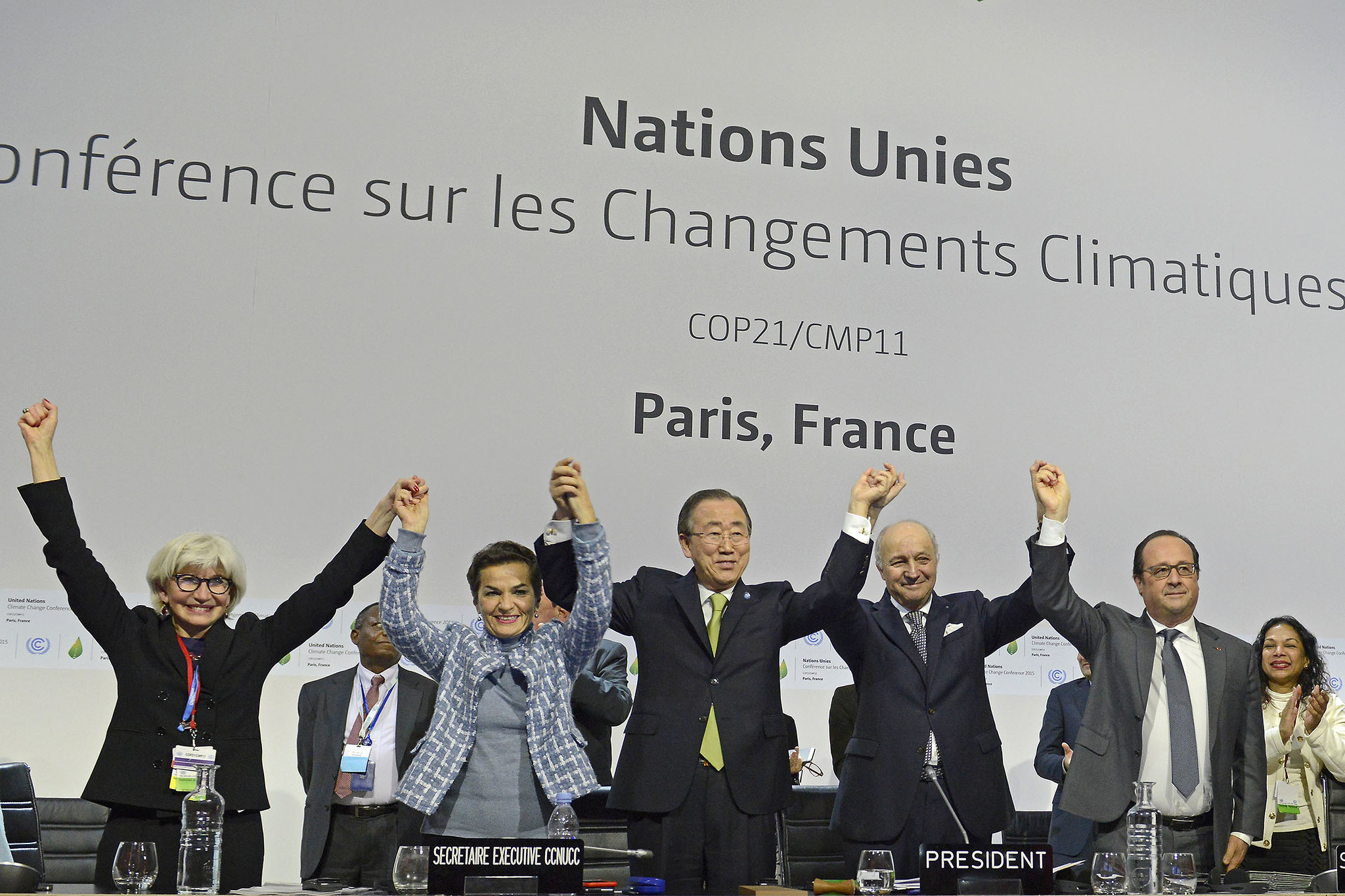 A imagem mostra cinco autoridades em um palco durante a 'COP21/CMP11' em Paris, França, com os braços levantados e as mãos dadas em um gesto de comemoração, diante de um painel branco com o texto 'Nations Unies Conférence sur les Changements Climatiques'. Da esquerda para a direita: Laurence Tubiana, uma mulher de pele branca, cabelo grisalho curto e óculos de armação escura, vestindo um blazer preto sobre uma blusa escura e um crachá; Christiana Figueres, de pele branca e cabelo castanho escuro curto, usando uma jaqueta estampada em azul e cinza; Ban Ki-moon, de ascendência asiática e cabelo preto, com terno escuro, camisa branca e gravata verde; Laurent Fabius, um homem de pele branca e cabelo branco, vestindo terno escuro, camisa branca e gravata escura estampada; e François Hollande, de pele branca, cabelo castanho e óculos, usando terno cinza, camisa branca e gravata escura.