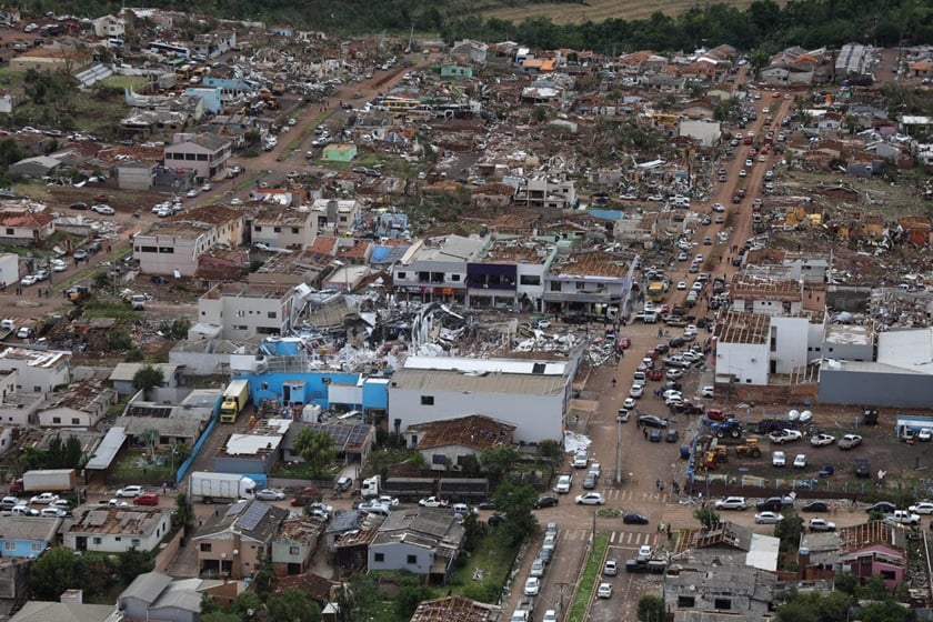A imagem é uma tomada aérea de uma área urbana que exibe danos extensos em múltiplas construções. Edifícios de poucos andares e casas estão com telhados total ou parcialmente destruídos, e escombros estão espalhados pelas propriedades e ruas. Uma via principal, visível no centro e à direita, está ladeada por numerosos veículos estacionados ou alinhados. Ao fundo, é possível ver uma área de vegetação verde e árvores.