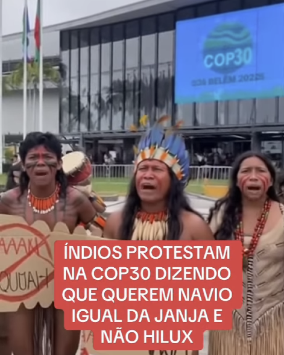 A imagem mostra três pessoas indígenas em pé, usando cocares, colares e pinturas corporais tradicionais. Duas delas seguram placas de protesto feitas de papelão. Ao fundo, aparece um prédio moderno com fachada de vidro e, acima dele, um grande painel azul onde se lê ‘COP30 – CIDA BELÉM 20225’. Também há mastros com bandeiras ao lado do prédio. Na frente das pessoas, um texto em letras brancas sob fundo vermelho, em destaque, diz: ‘ÍNDIOS PROTESTAM NA COP30 DIZENDO QUE QUEREM NAVIO IGUAL DA JANJA E NÃO HILUX’.