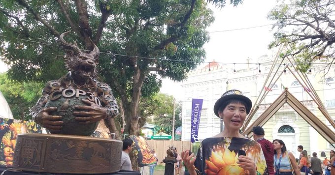 A imagem mostra uma cena ao ar livre, com árvores e pessoas ao fundo. À esquerda, há uma escultura de aparência fantástica posicionada sobre um pedestal decorado; a figura segura um globo com a inscrição ‘COP30’. À direita, está uma pessoa sorrindo e falando, vestindo uma camisa estampada com uma grande flor alaranjada e usando um chapéu. Atrás dela, aparecem outras pessoas, banners verticais e estruturas de madeira em formato triangular. Ao fundo, vê-se parte de um prédio claro com janelas arqueadas.