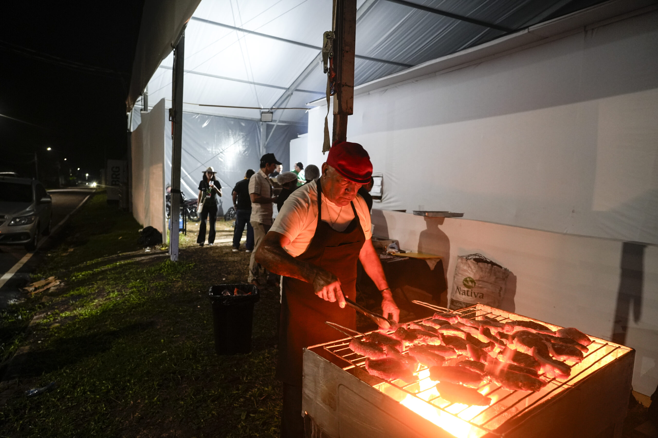 A imagem mostra um homem usando boné vermelho, camiseta clara e avental escuro, assando pedaços de carne em uma churrasqueira iluminada pelo fogo em uma área externa coberta por uma estrutura de lona branca durante a noite, enquanto ao fundo outras pessoas circulam e conversam perto de mesas e equipamentos sob a mesma cobertura.