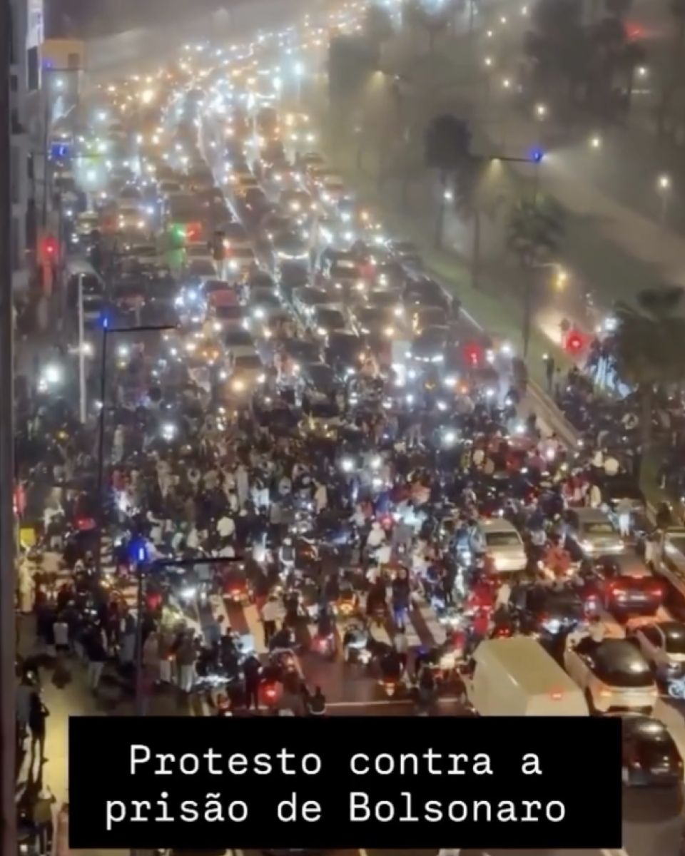 Print de publicação no Instagram mostra uma grande concentração de pessoas e veículos ocupando múltiplas faixas de uma avenida iluminada, durante a noite, com muitos faróis e luzes de celulares acesos. Na parte inferior há um retângulo preto com o texto ‘Protesto contra a prisão de Bolsonaro’.