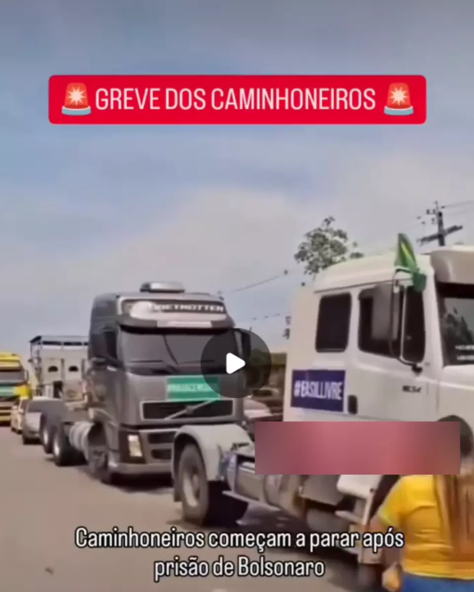 A imagem mostra uma fila de caminhões estacionados em uma via, incluindo veículos de cores claras e escuras, alguns com adesivos e uma bandeira do Brasil fixada na lateral de um deles; em primeiro plano, uma pessoa usando camiseta amarela e shorts azuis caminha pela rua. Na parte superior aparece o texto ‘GREVE DOS CAMINHONEIROS’, e na parte inferior lê-se ‘Caminhoneiros começam a parar após prisão de Bolsonaro’.