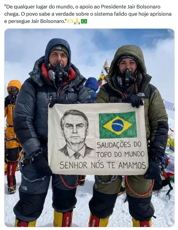  A imagem mostra duas pessoas vestidas com roupas grossas de montanhismo, incluindo jaquetas acolchoadas, luvas escuras, calças com proteções térmicas e máscaras de oxigênio, em um ambiente de neve; elas seguram um pano retangular que exibe um desenho do rosto de um homem e a bandeira do Brasil, acompanhado do texto ‘Saudações do topo do mundo senhor nós te amamos’, enquanto outras pessoas com vestimentas semelhantes aparecem ao fundo.