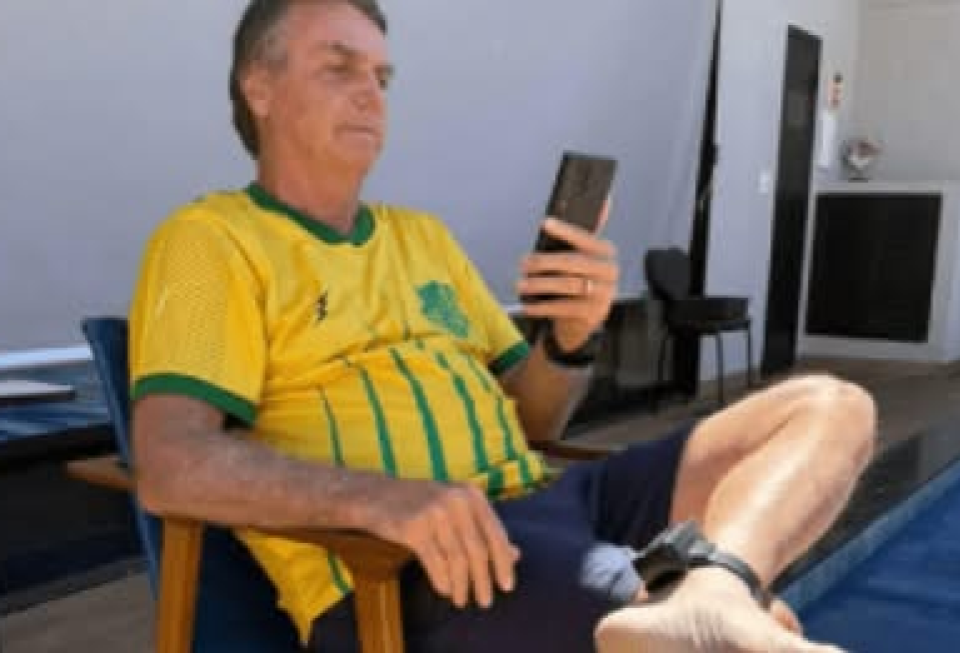 A imagem é uma captura de tela de uma publicação em rede social que apresenta texto sobre alegações envolvendo a tornozeleira eletrônica usada por Jair Bolsonaro e contaminação por césio-137; logo abaixo, há uma fotografia que mostra Bolsonaro — homem de pele clara que aparece vestindo camisa nas cores verde e amarelo, sentado em uma cadeira ao ar livre, com uma tornozeleira eletrônica presa à perna esquerda e segurando um celular na mão direita — próximo a uma área com piscina e móveis externos.
