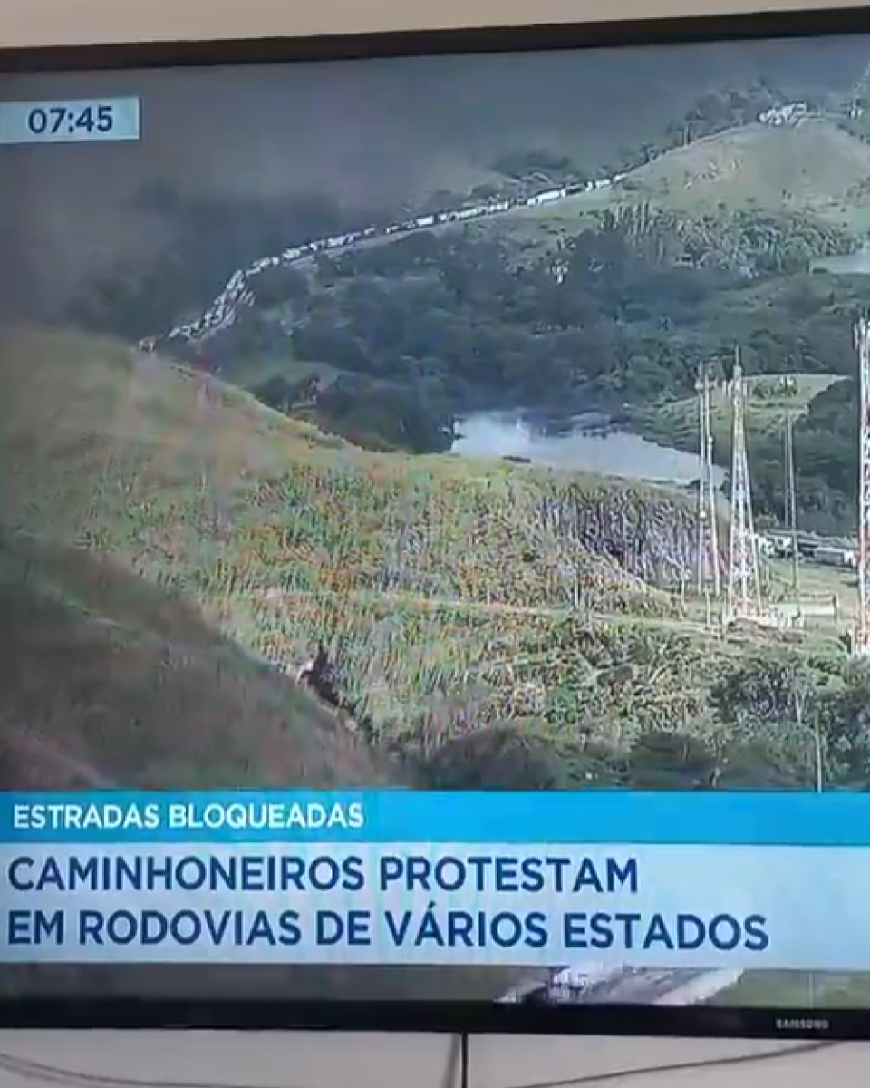 A imagem mostra a tela de um televisor exibindo uma transmissão jornalística com a paisagem de uma rodovia cercada por morros e vegetação, onde se observa uma longa fila de veículos; na parte superior esquerda há um marcador de horário com os números ‘07:45’, e na faixa inferior aparecem as legendas ‘ESTRADAS BLOQUEADAS’ e ‘CAMINHONEIROS PROTESTAM EM RODOVIAS DE VÁRIOS ESTADOS’.