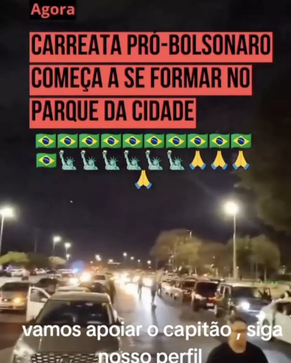 A imagem mostra uma via urbana à noite com várias fileiras de carros parados ou em movimento lento, alguns com faróis acesos; há pessoas caminhando entre os veículos e árvores visíveis ao fundo sob iluminação pública. Textos sobrepostos incluem a frase ‘Agora Carreata pró-Bolsonaro começa a se formar no Parque da Cidade’, acompanhada de emojis de bandeiras, estátuas e mãos em gesto de oração, além de outra frase parcial na parte inferior.