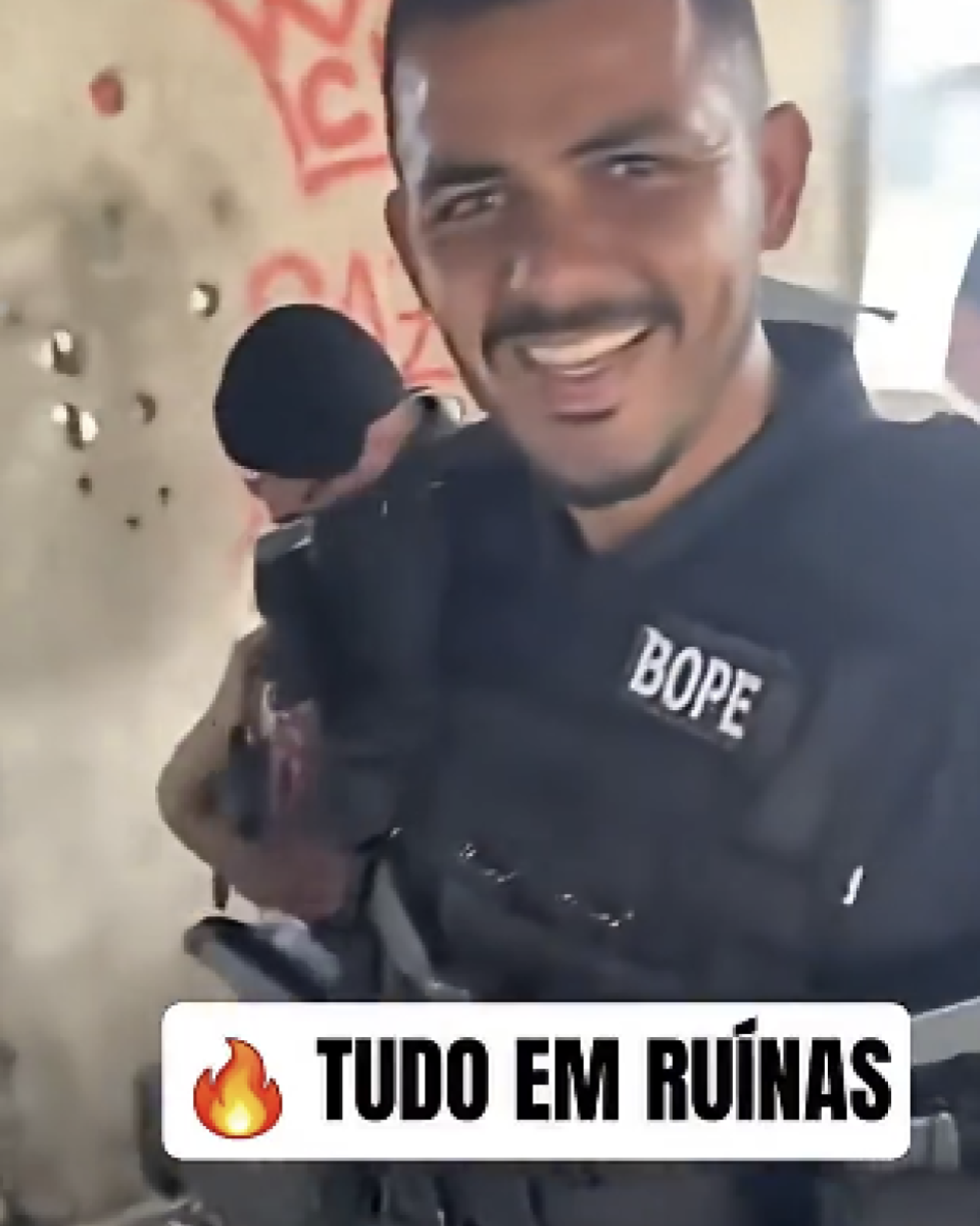 A imagem mostra um homem sorrindo, vestindo um colete tático preto com a palavra ‘BOPE’ escrita em branco na parte frontal. Ele carrega um equipamento preso ao corpo e está em um ambiente interno com paredes desgastadas e pichações ao fundo. À esquerda, parcialmente visível, há outra pessoa usando gorro preto e também equipada, posicionada atrás dele. Na parte inferior da imagem aparece a frase ‘TUDO EM RUÍNAS’.