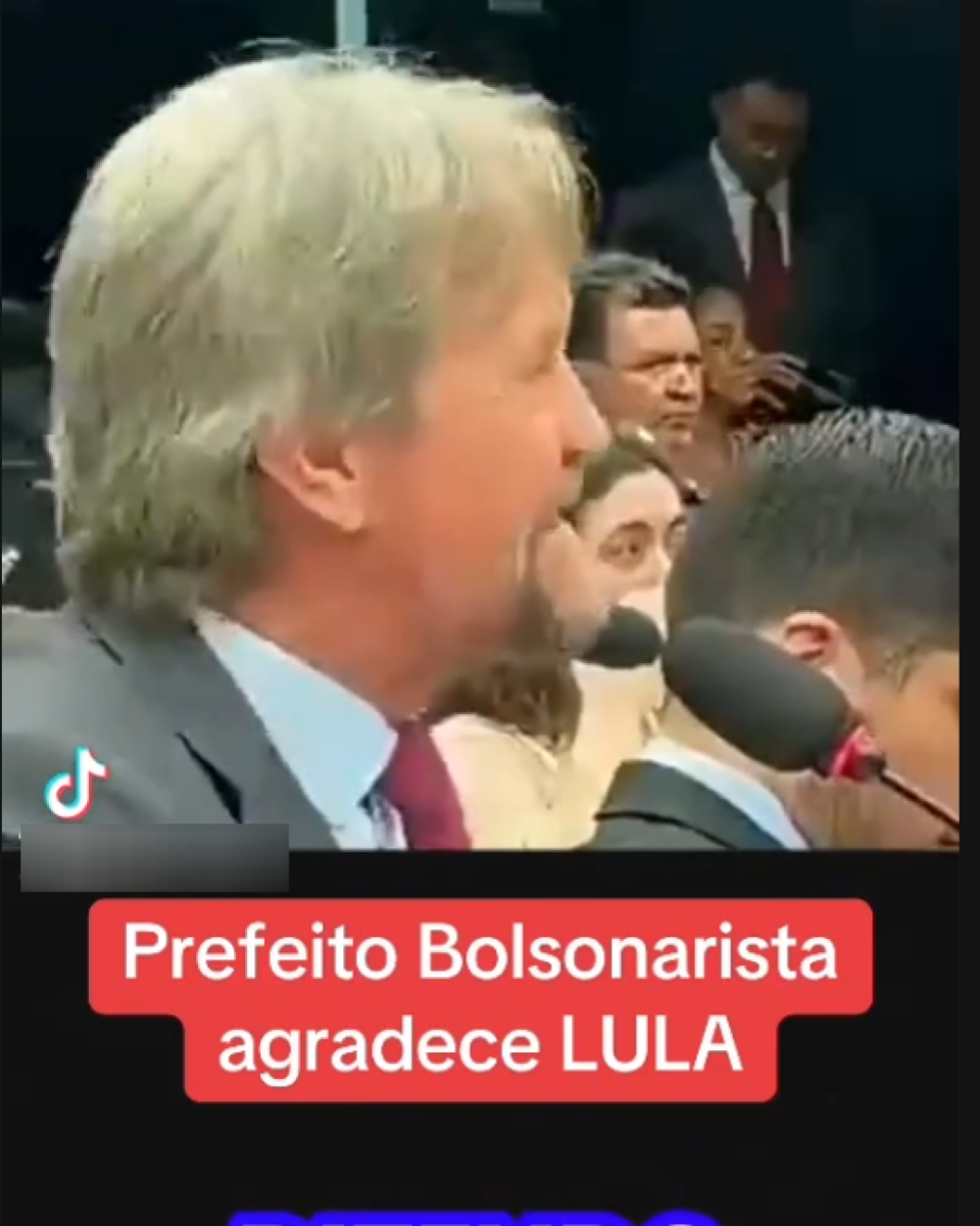 A imagem mostra um homem de pele clara, cabelos grisalhos e barba curta, usando terno cinza e gravata bordô, falando ao microfone em um ambiente semelhante a uma audiência ou sessão formal, com outras pessoas sentadas ao fundo. No canto inferior esquerdo, há o logotipo do TikTok. Sobre a imagem, há textos em destaque que dizem: ‘Prefeito Bolsonarista agradece LULA’.