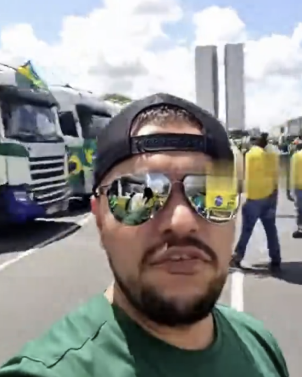 A imagem mostra um homem em primeiro plano, usando boné preto para trás, óculos espelhados e camisa verde. Ele está segurando a câmera ou celular apontado para si, enquadrando o próprio rosto. No reflexo dos óculos, é possível ver pessoas andando pela rua e veículos próximos. Próximo aos óculos também é possível ver um borrão na imagem. Atrás dele, há várias carretas brancas com faixas ou pinturas nas cores verde e amarelo, com bandeiras do Brasil presas a algumas delas. Ao fundo também é possível ver pessoas vestindo roupas nas cores da bandeira brasileira e, mais distante, duas torres retangulares altas lado a lado, sob um céu parcialmente nublado.