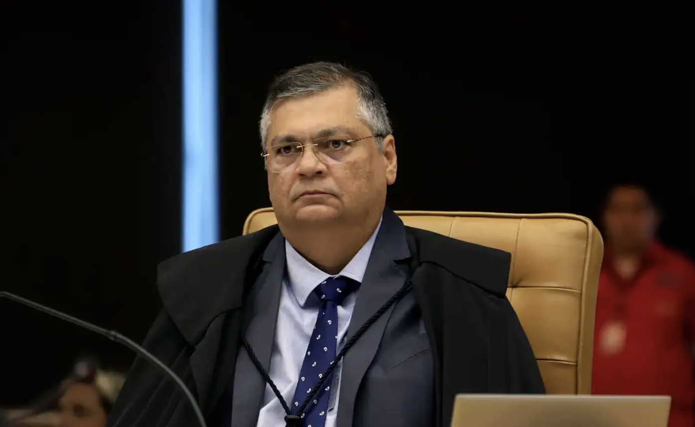 A imagem mostra um o ministro Flávio Dino sentado em uma cadeira de encosto alto de cor bege, usando óculos, terno escuro, camisa clara e gravata azul com pequenos detalhes. Ele veste também uma capa preta sobre o terno. À frente dele há um microfone e a lateral de um notebook visível no canto inferior direito. O fundo é escuro, com uma pessoa desfocada ao fundo, vestindo roupa vermelha.