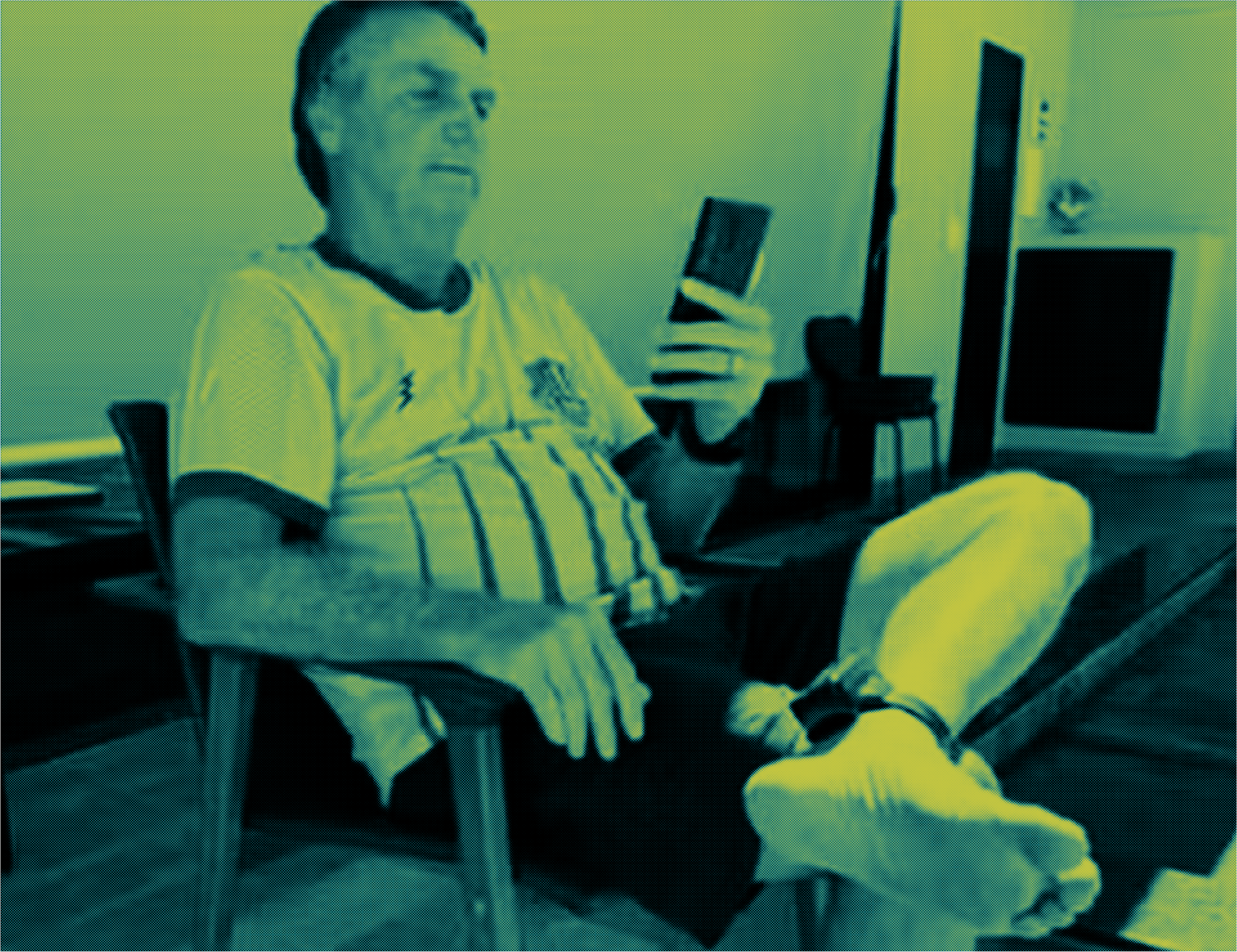 Foto mostra Bolsonaro, um homem branco, sentado com as pernas cruzadas em uma cadeira. Na perna esquerda, é possível ver a tornozeleira eletrônica. Ele sorri enquanto olha a tela do celular em sua mão esquerda.